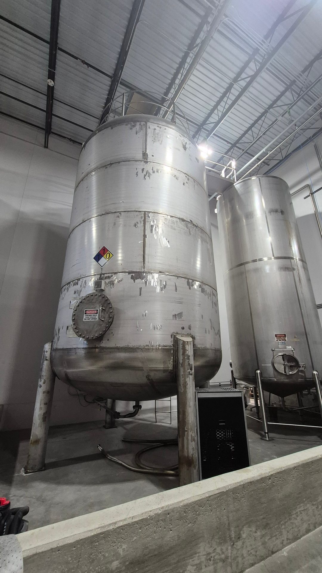 Two large, stainless steel industrial tanks with an industrial ceiling structure in a factory setting.