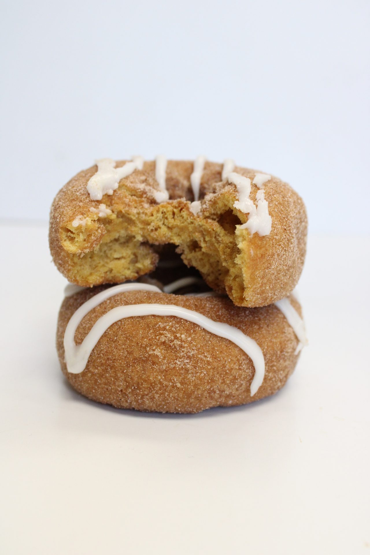 Two cinnamon sugar donuts with white icing, top one has a bite taken out, stacked on a white surface.