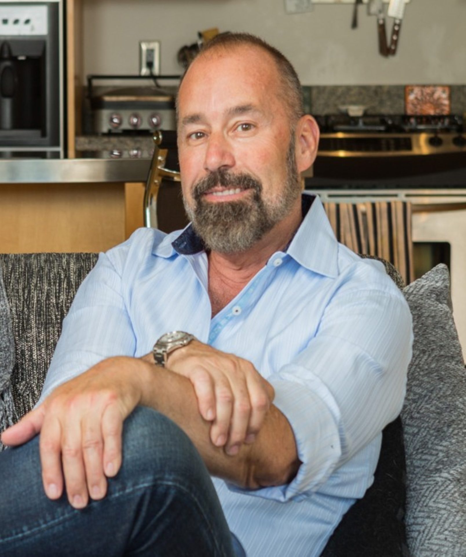 A smiling man with a beard and light blue shirt sits on a couch with crossed hands.