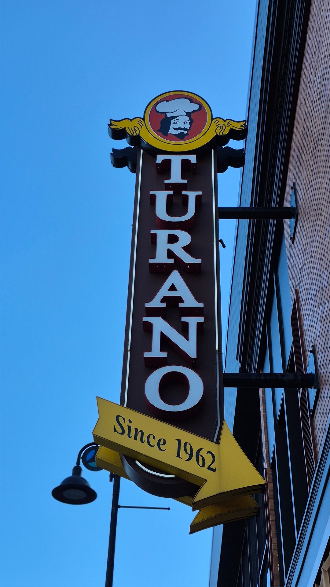 Vertical "TURANO" sign with chef logo, "Since 1962" on an arrow, against a blue sky.