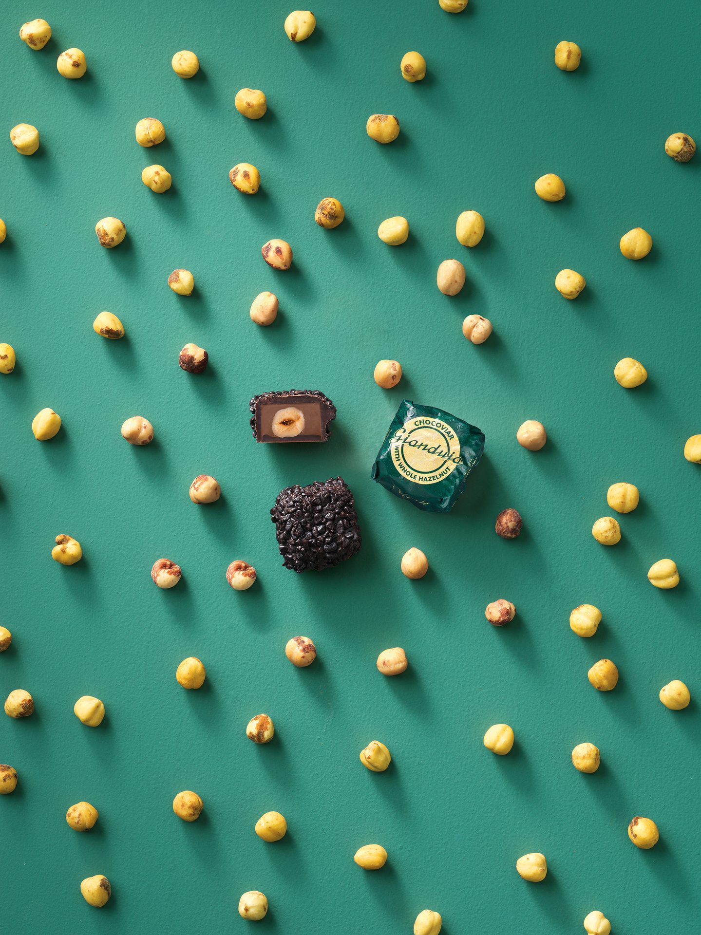 Scattered hazelnuts on green, framing a wrapped, an unwrapped hazelnut chocolate, and dark chocolate.