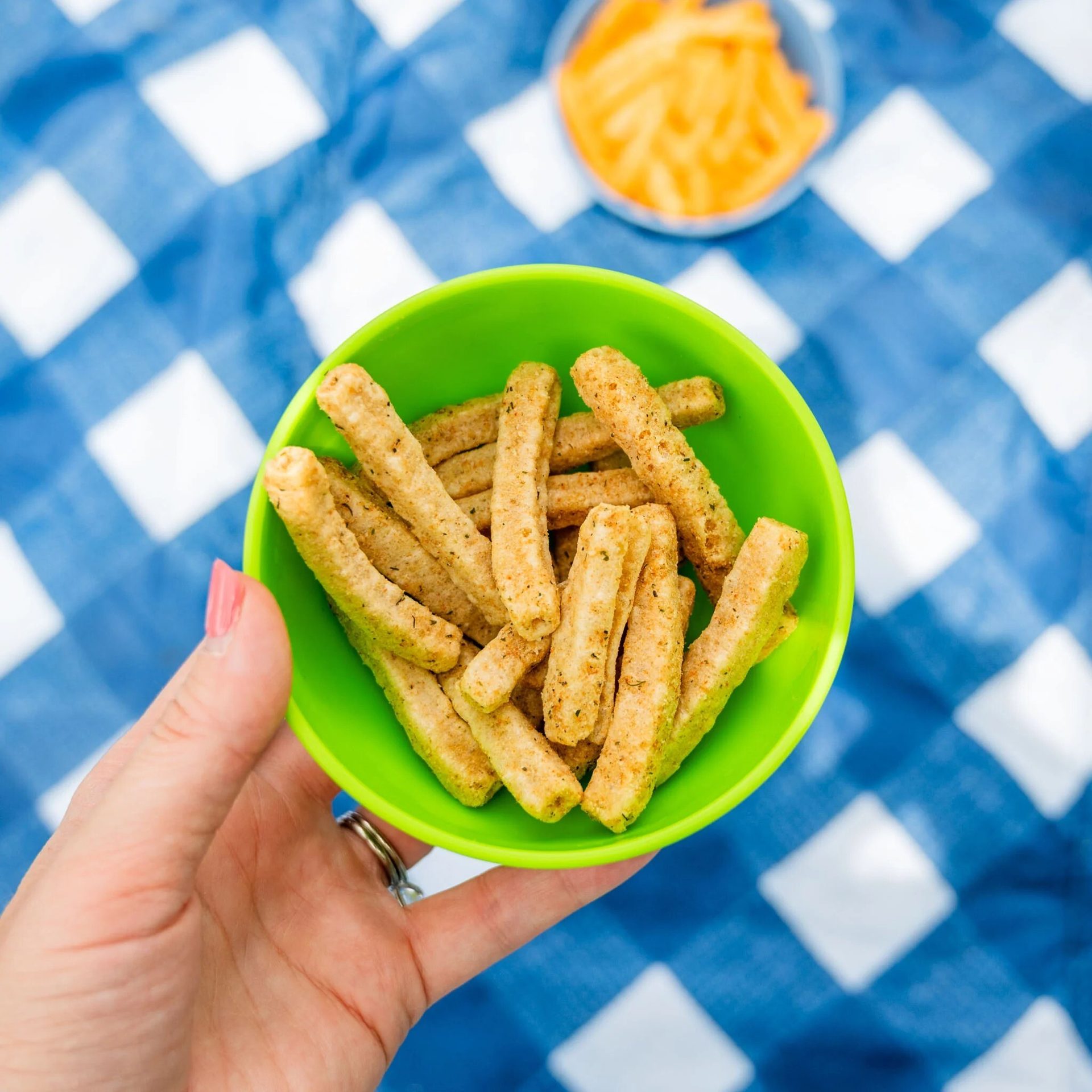 A hand with pink nail polish holds a green bowl of savory snack sticks on a blue and white checkered tablecloth.