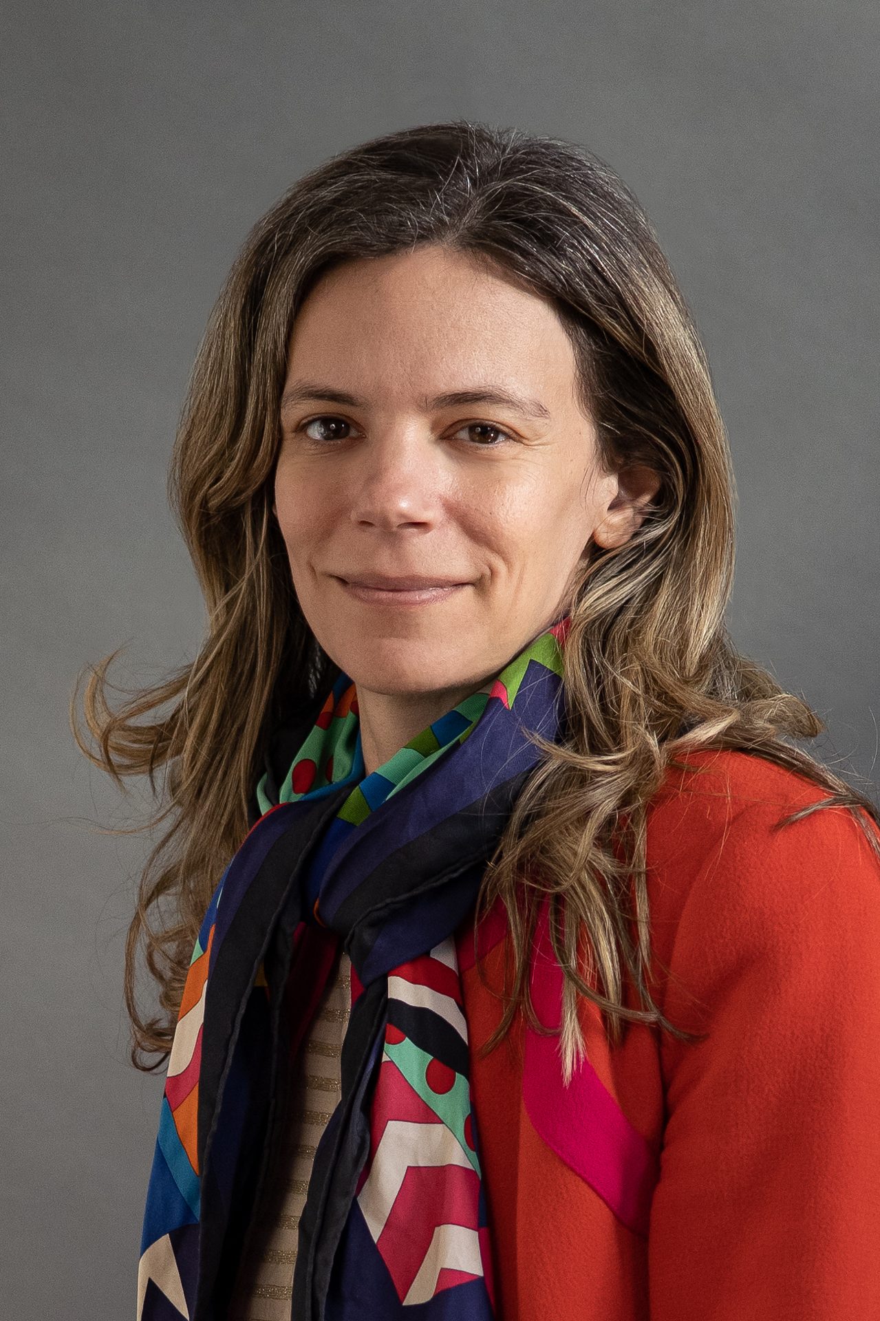 Smiling woman in red jacket and colorful scarf with long brown hair.