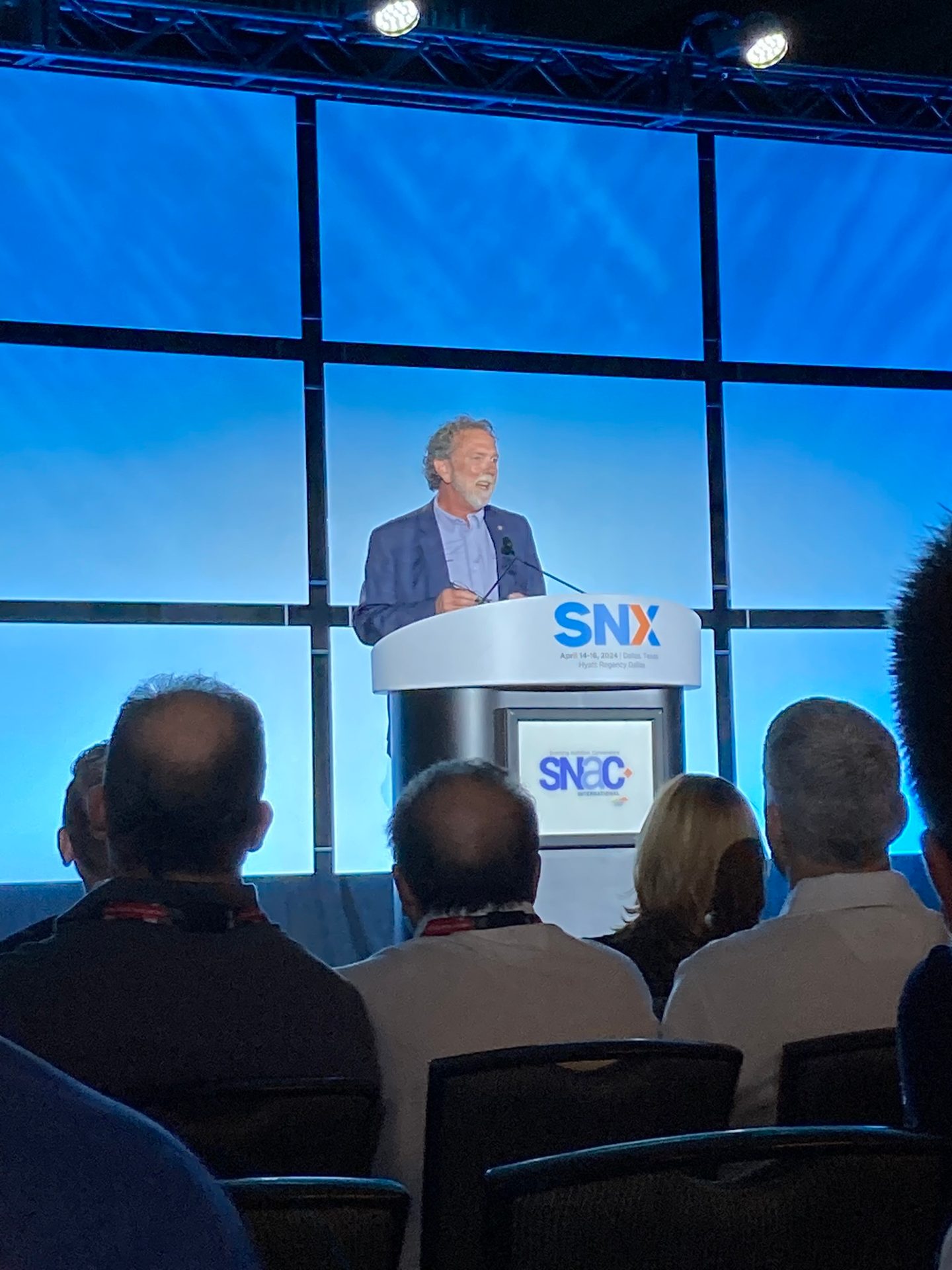 Man speaking at a conference podium with SNX branding, an audience seated in the foreground.