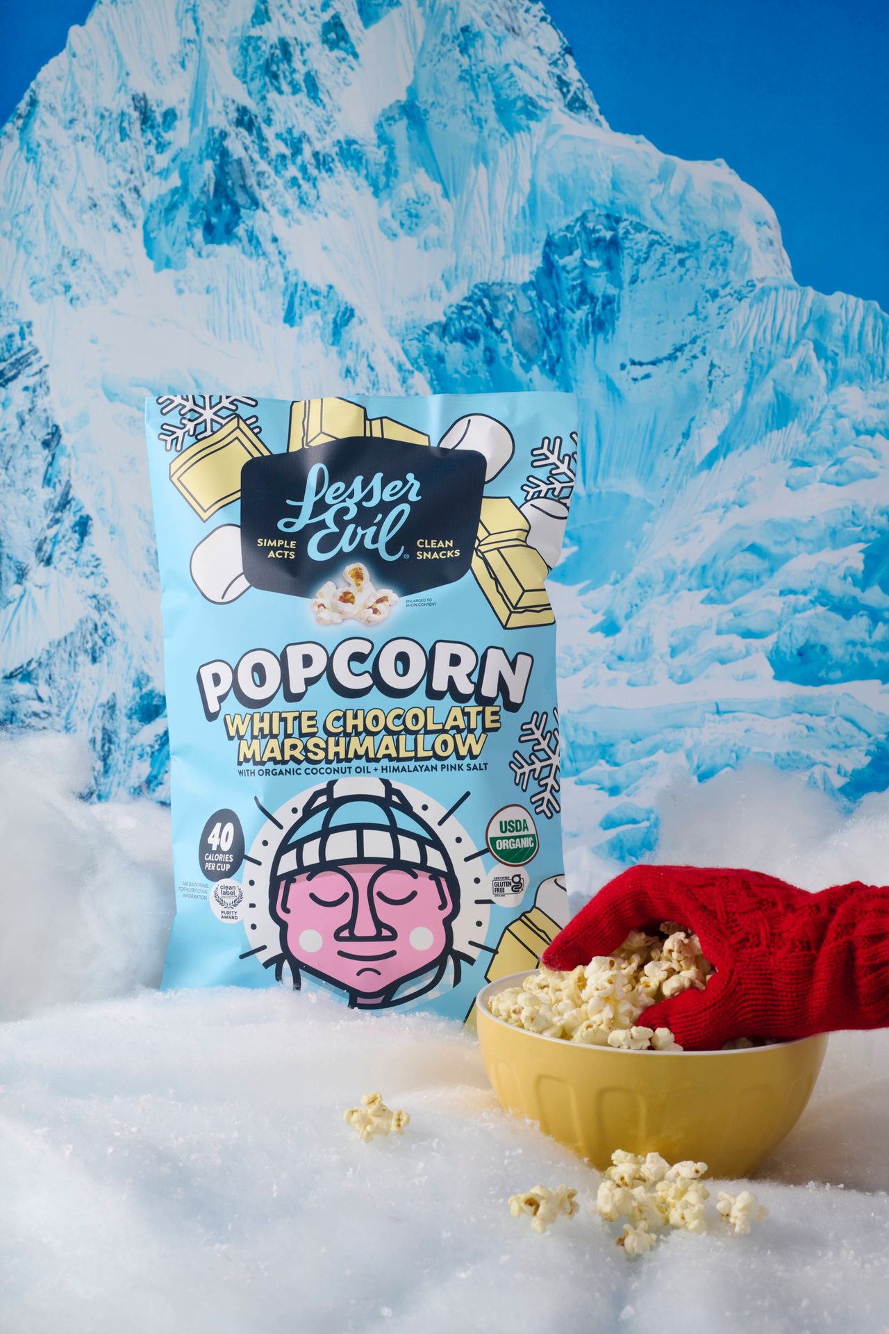 Bag of White Chocolate Marshmallow popcorn in snow. Hand in red mitten grabs popcorn from a yellow bowl.