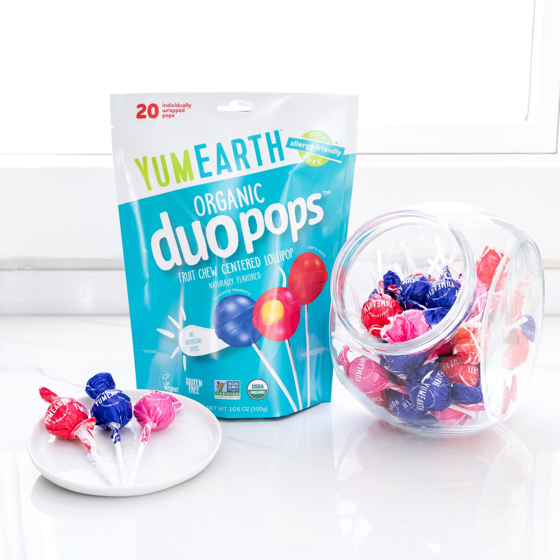 YumEarth Organic Duo Pops in blue packaging, surrounded by a candy jar full of the lollipops and a tray with three lollipops on it.