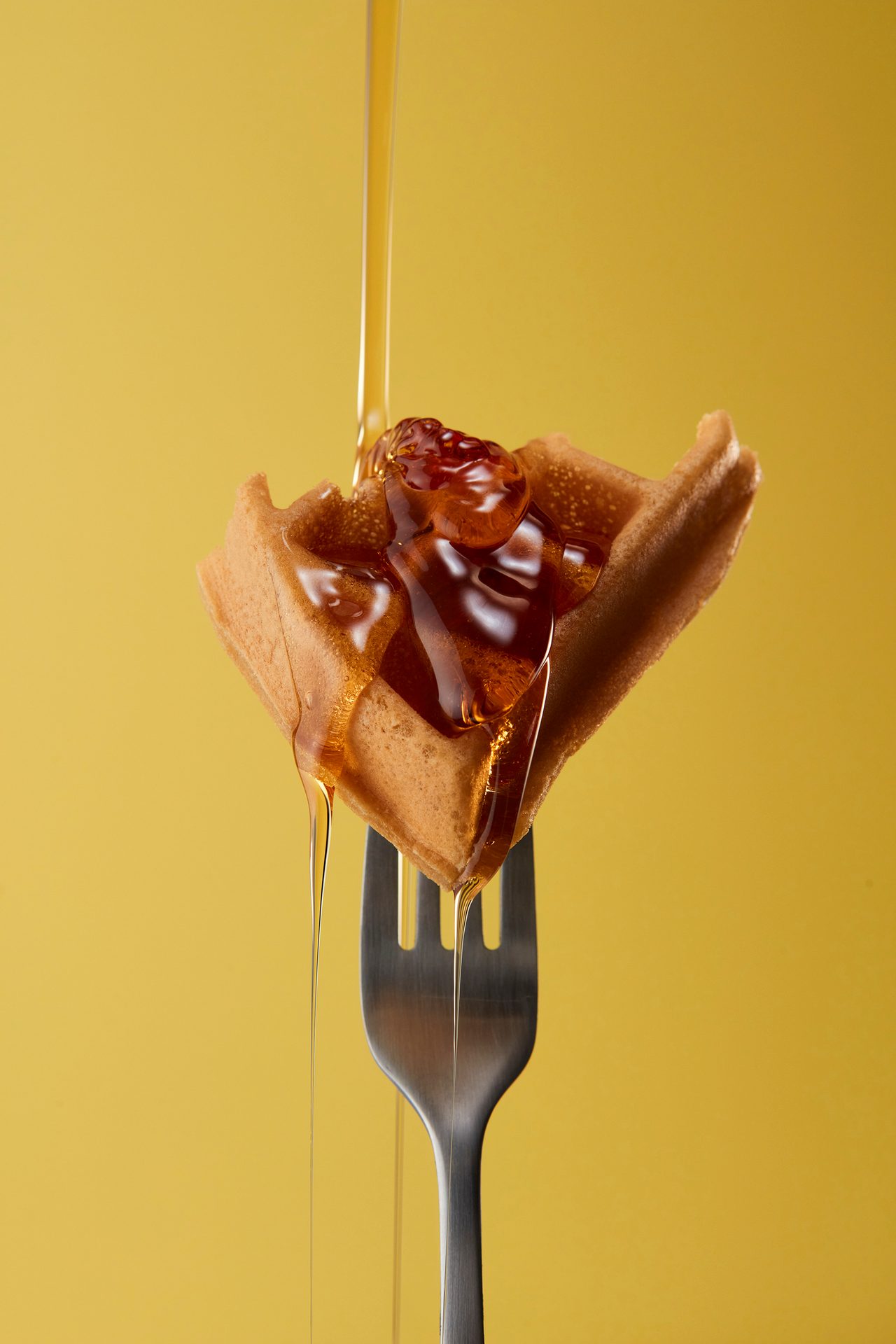 A golden waffle on a fork with honey pouring over it and a berry topping, against a yellow background.