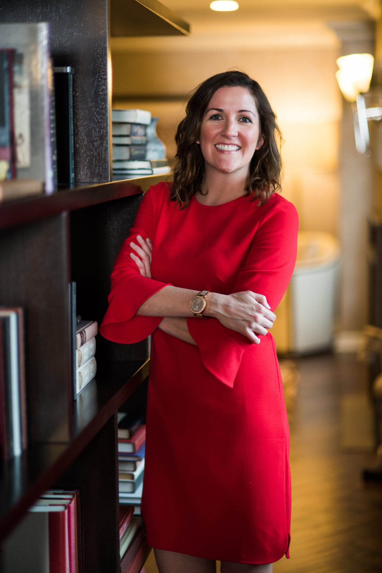 A happy woman in a red dress with crossed arms stands beside a dark bookshelf, smiling.