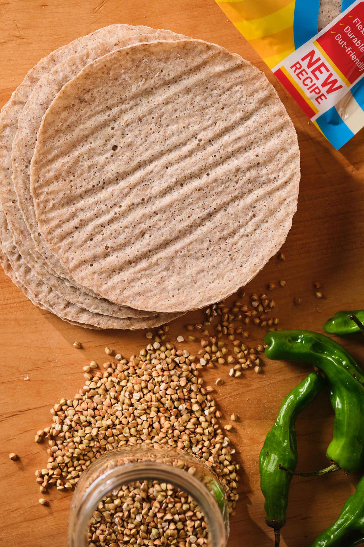 Stack of buckwheat tortillas, raw buckwheat, and green peppers on a wooden board with packaging.