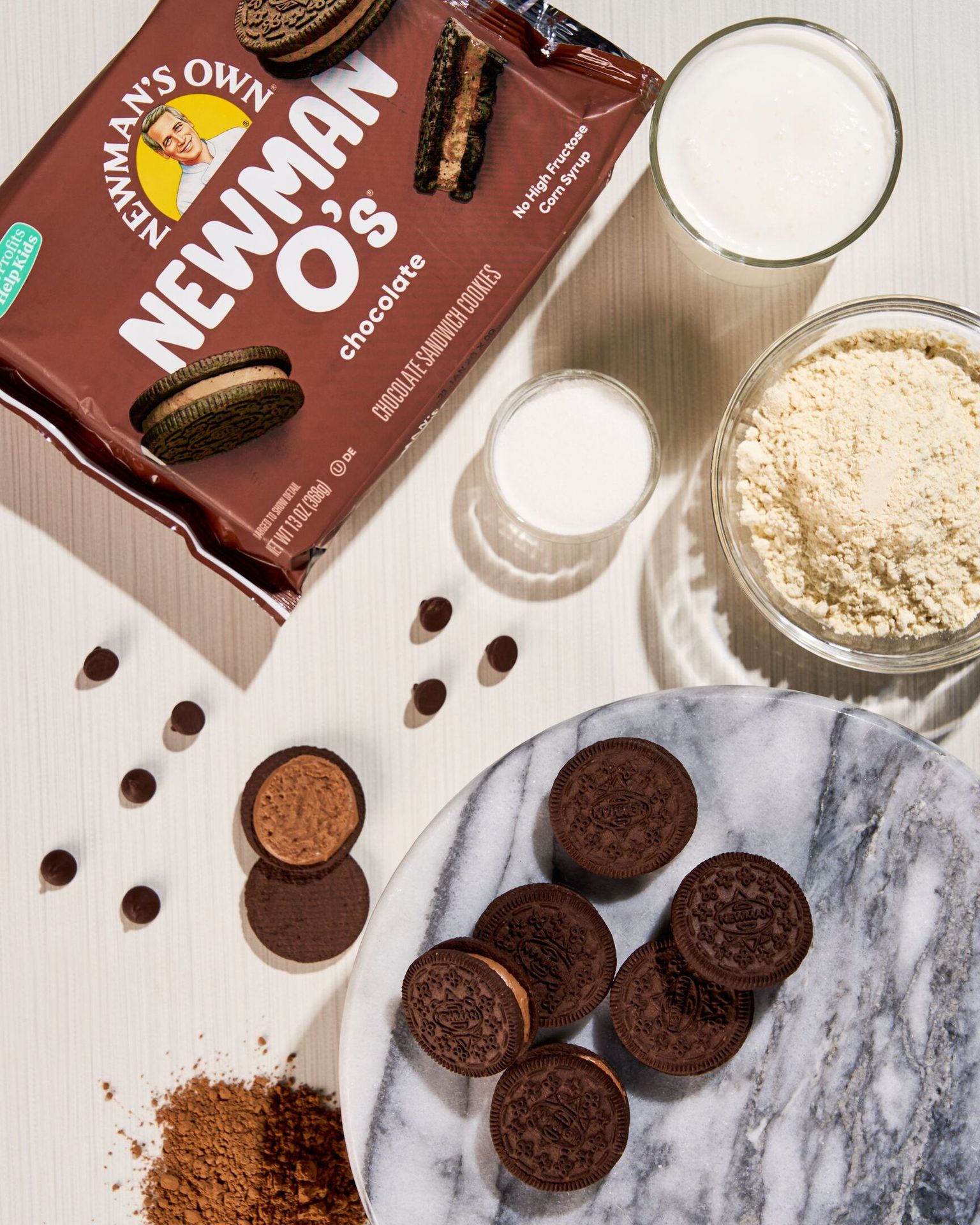 Newman's Own chocolate sandwich cookies, milk, and baking ingredients on a light surface.
