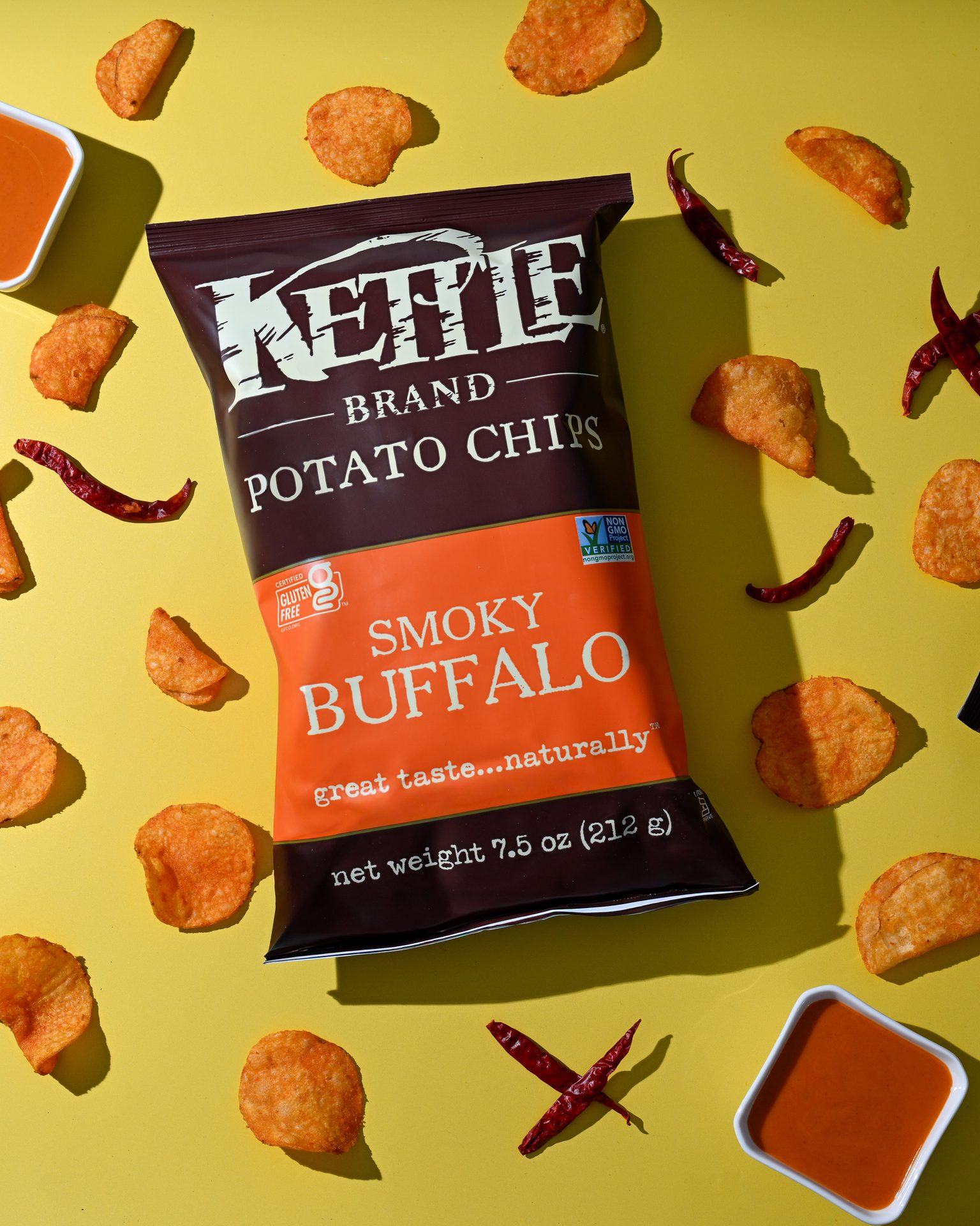 Bag of Kettle Brand Smoky Buffalo potato chips surrounded by loose chips, dried chilies, and dip.