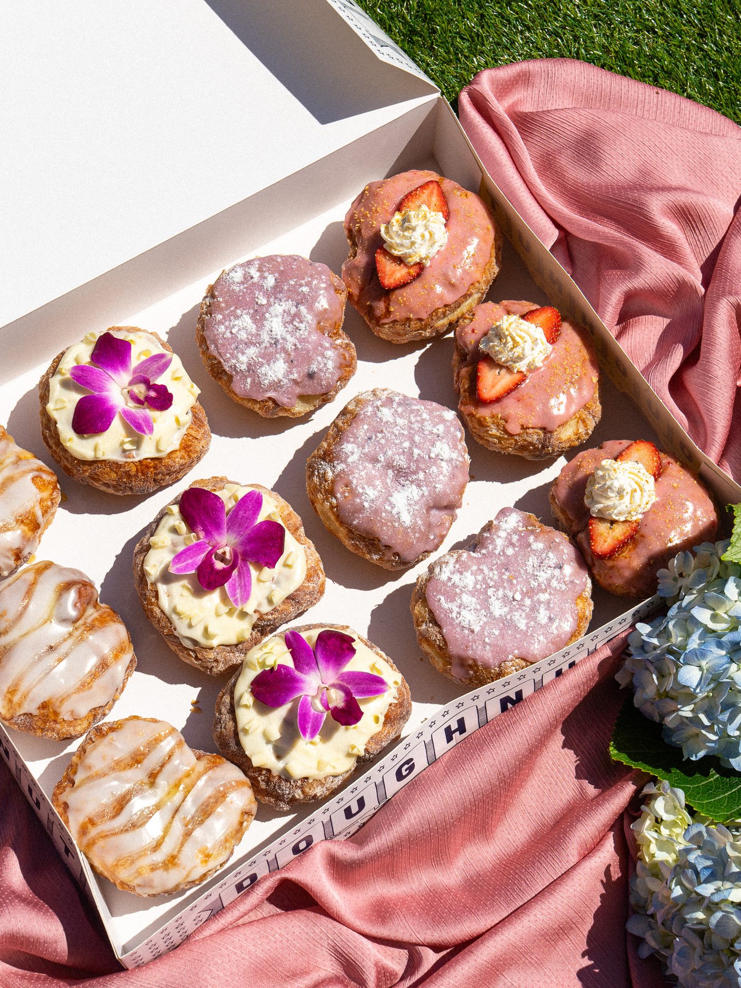 A box of assorted gourmet donuts with various glazes and toppings, including strawberries and orchids.
