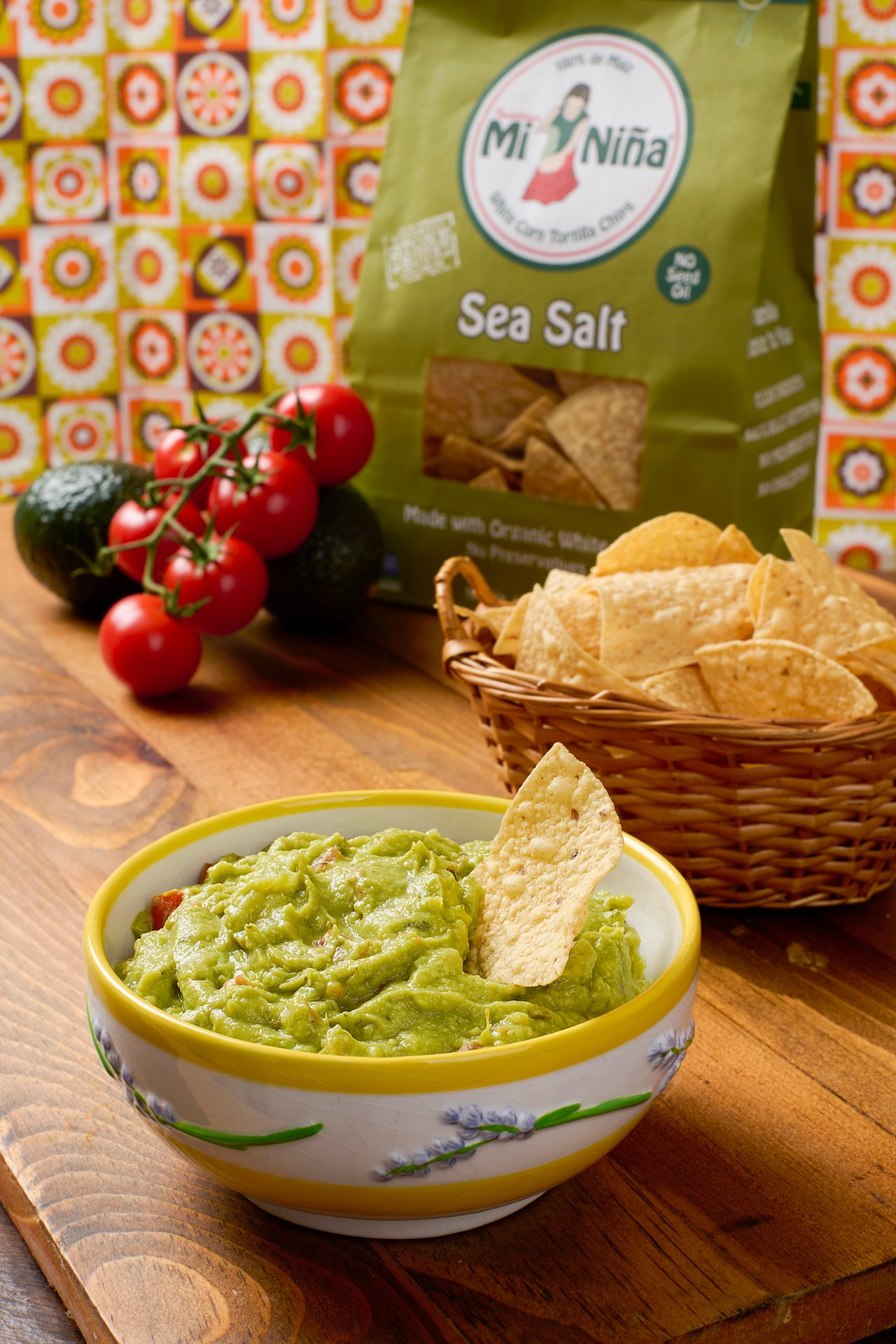 A bowl of guacamole with a chip, Mi Niña Sea Salt tortilla chips, fresh tomatoes, and avocados on wood.