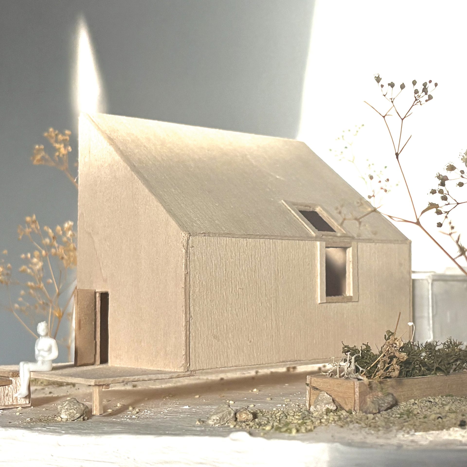 Light wooden architectural model of a house with a sloped roof, small figure, and dried plants.