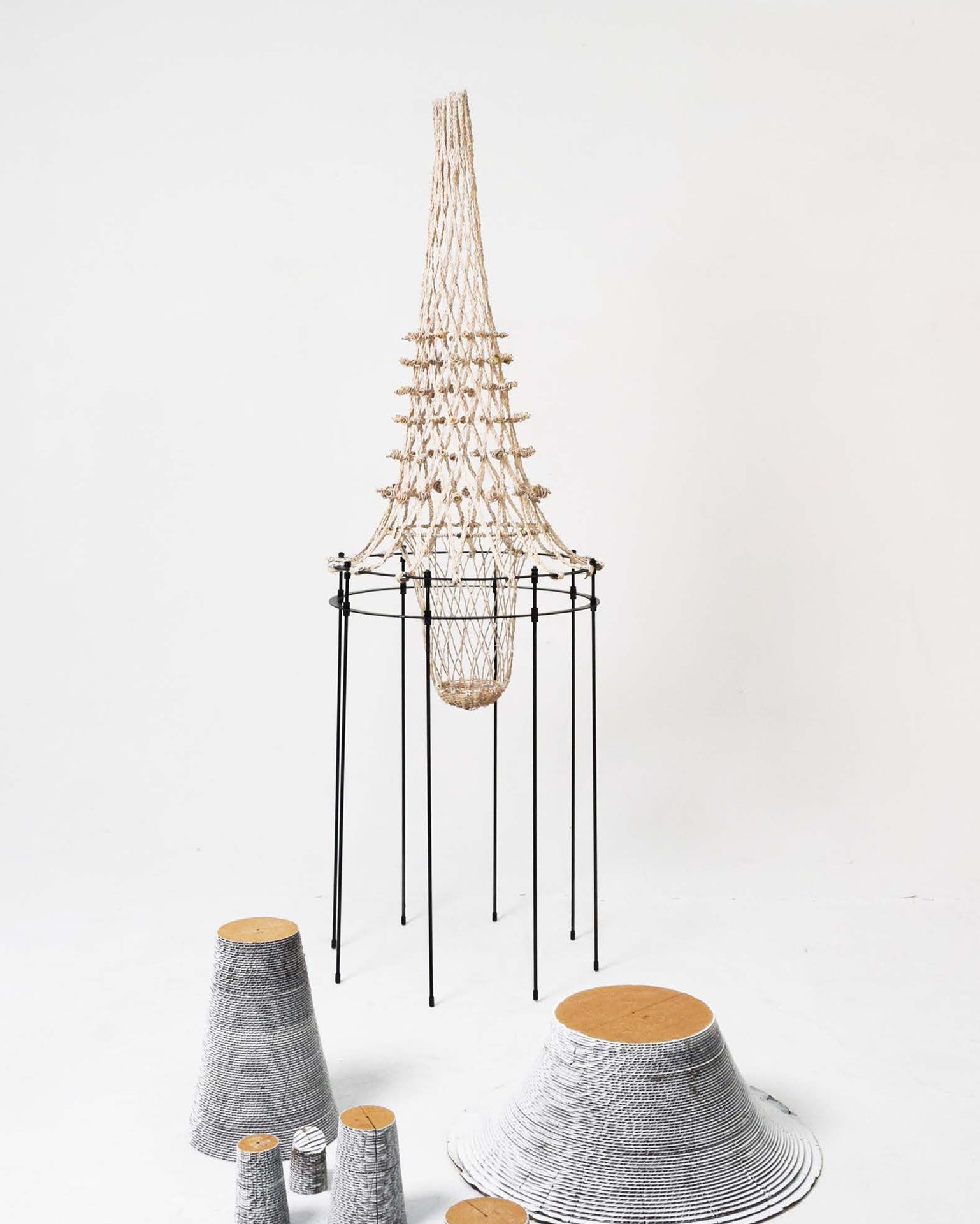 Woven bell-shaped form on black stand, with conical ridged objects, white background.