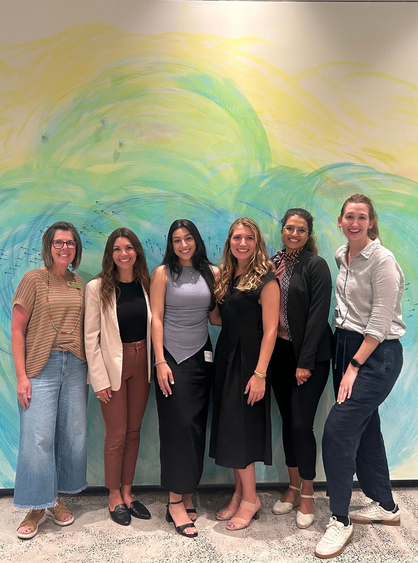 Six women smiling in front of a vibrant, swirly yellow, green, and blue mural.