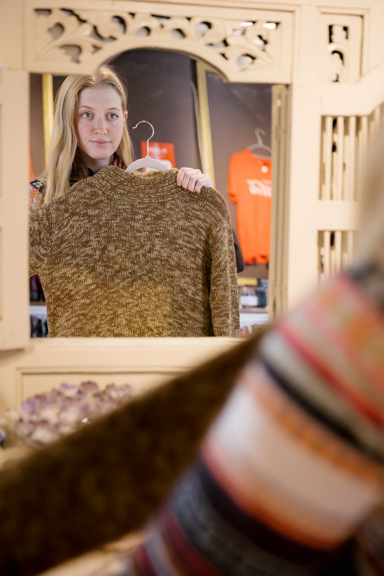en kvinne med blondt, langt h&#xE5;r som holder opp en brun strikket genser foran et speil mens hun ser p&#xE5; den i en bruktbutikk p&#xE5; &#xC5;rnes.
