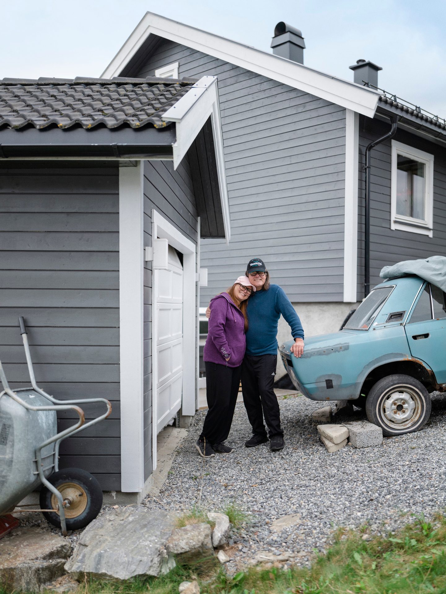 Kvinne og mann st&#xE5;r lent inntil hverandre ved en eldre bil, ved en garasje og foran et hus
