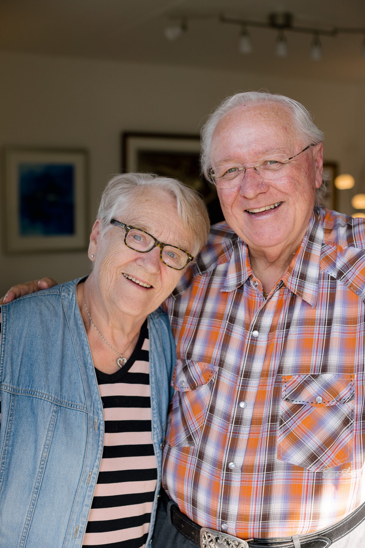 et eldre par med hvitt h&#xE5;r, briller og m&#xF8;nstret skjorte st&#xE5;r sammen og smiler inn i kameraet. 