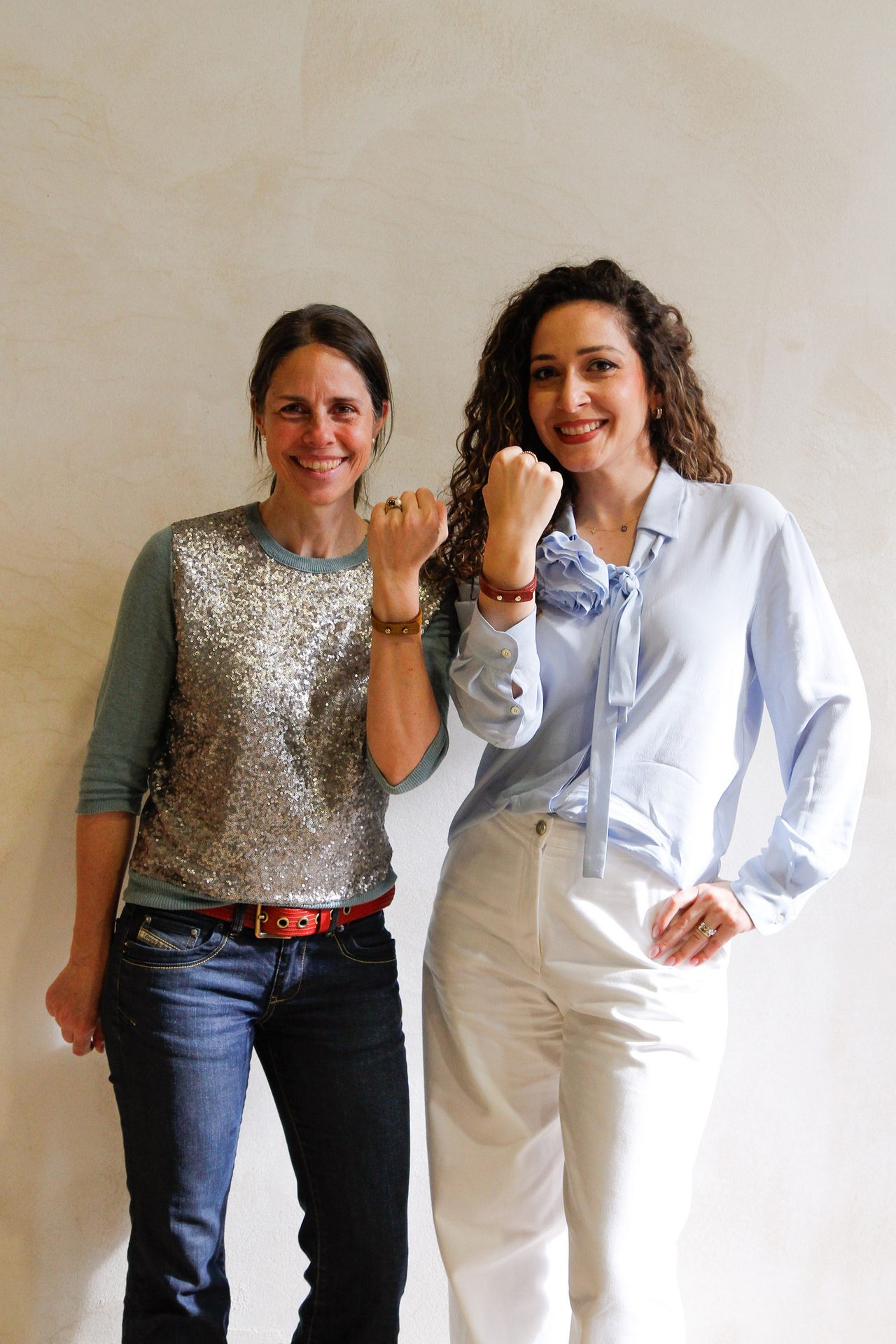Two smiling women in casual attire, one in sequins, the other in a blouse, both show fisted hands.