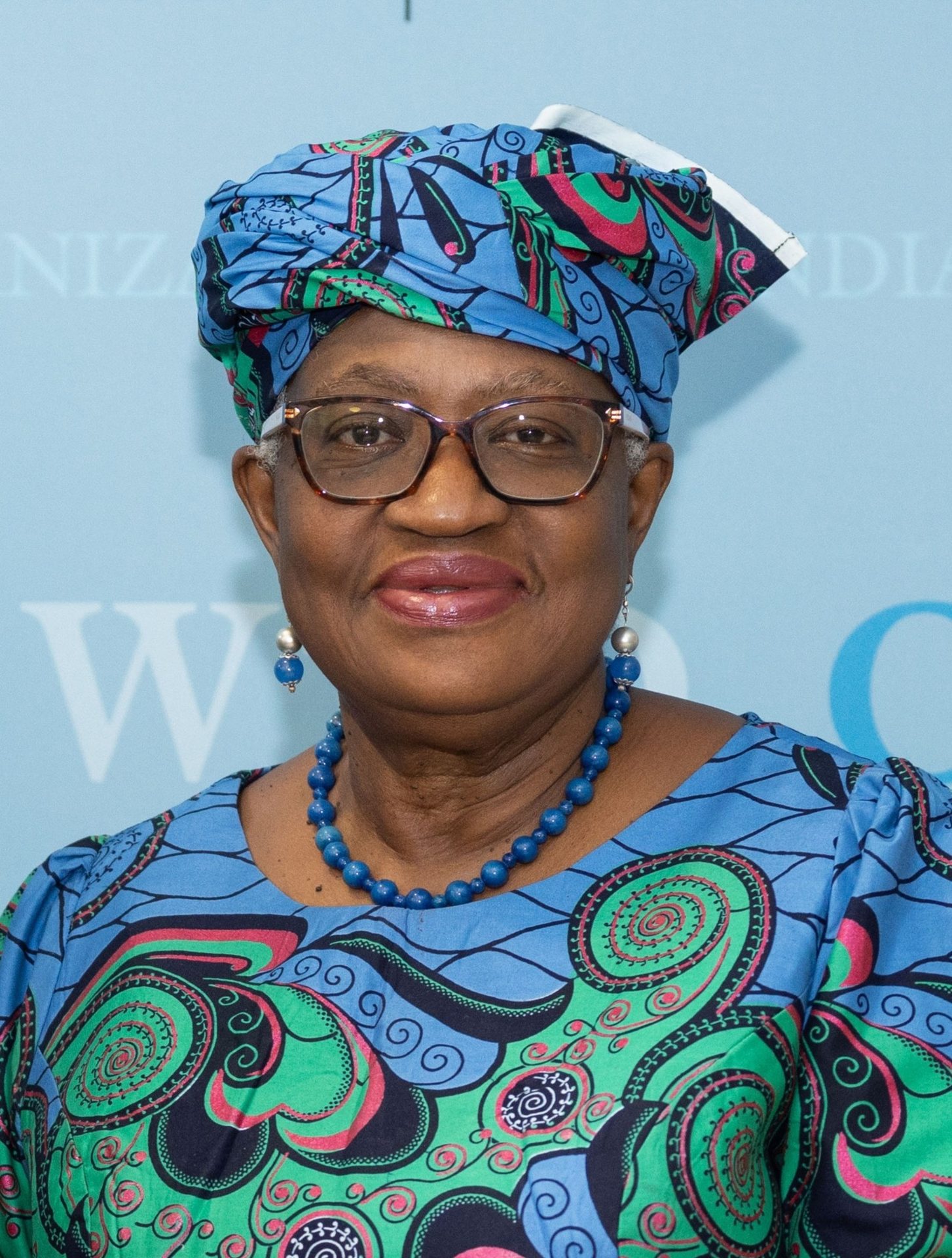 A woman in glasses and a vibrant patterned headwrap, wearing a matching dress, smiling.