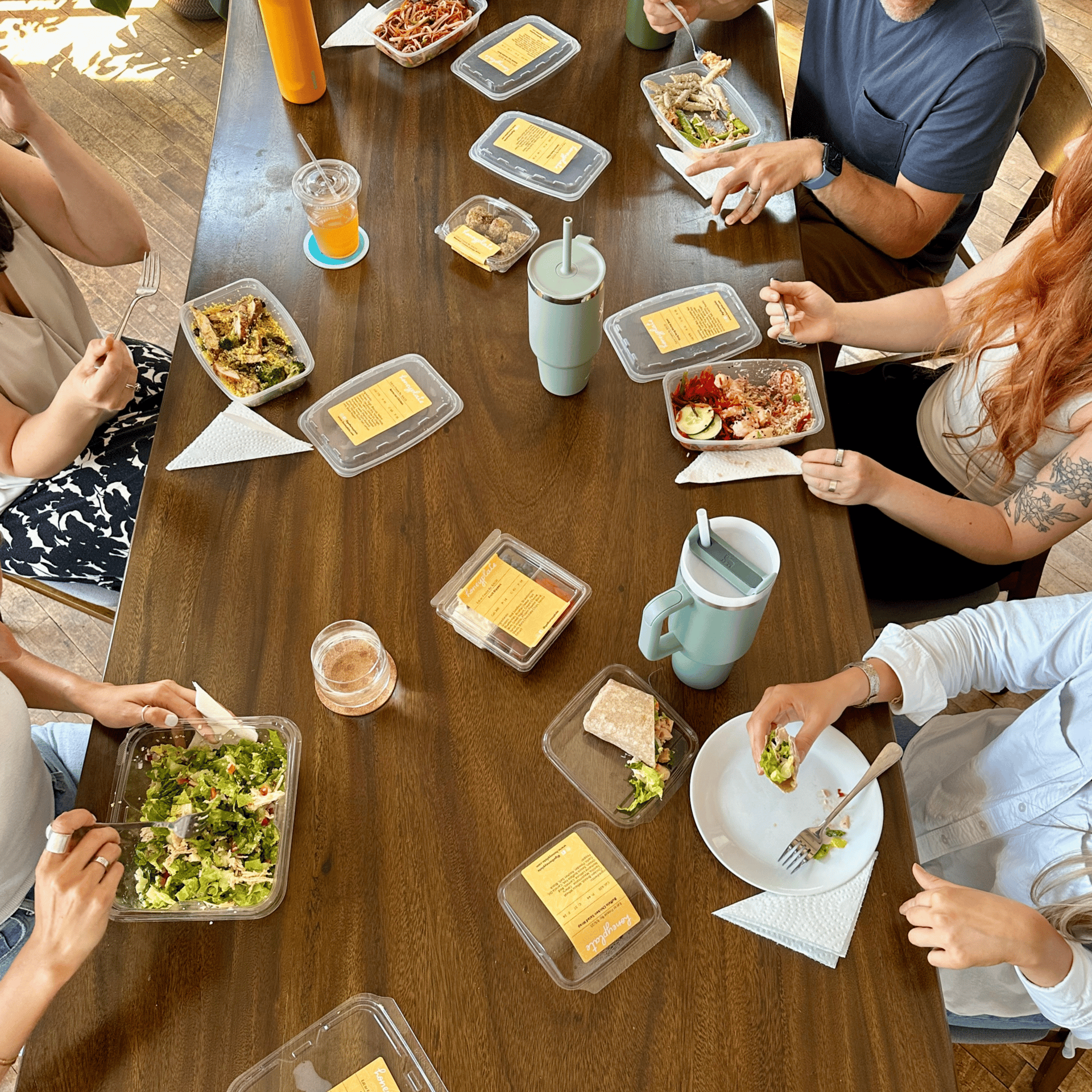 People gathered around a wooden table, eating various meals from transparent containers and cups.