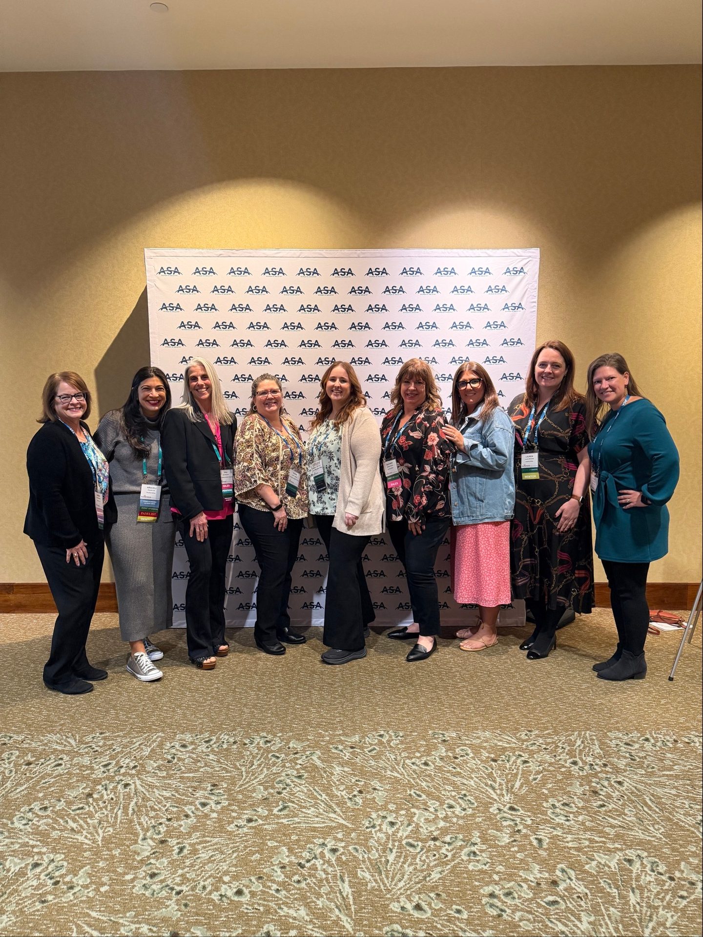 Nine women in professional attire smile, standing in a line before a white banner with ASA logos.