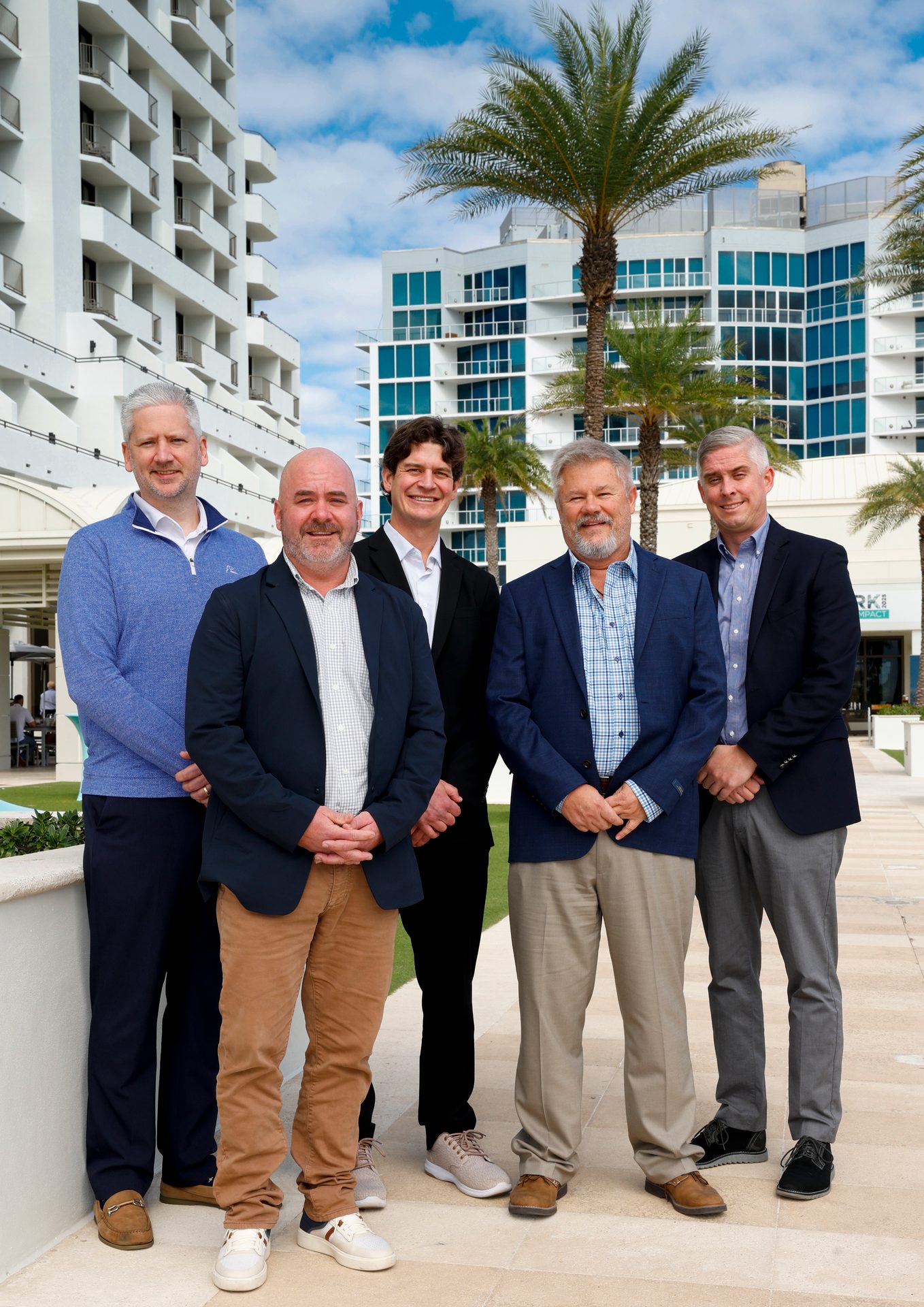 Five smiling men in business casual attire standing outdoors in front of white buildings and palm trees.