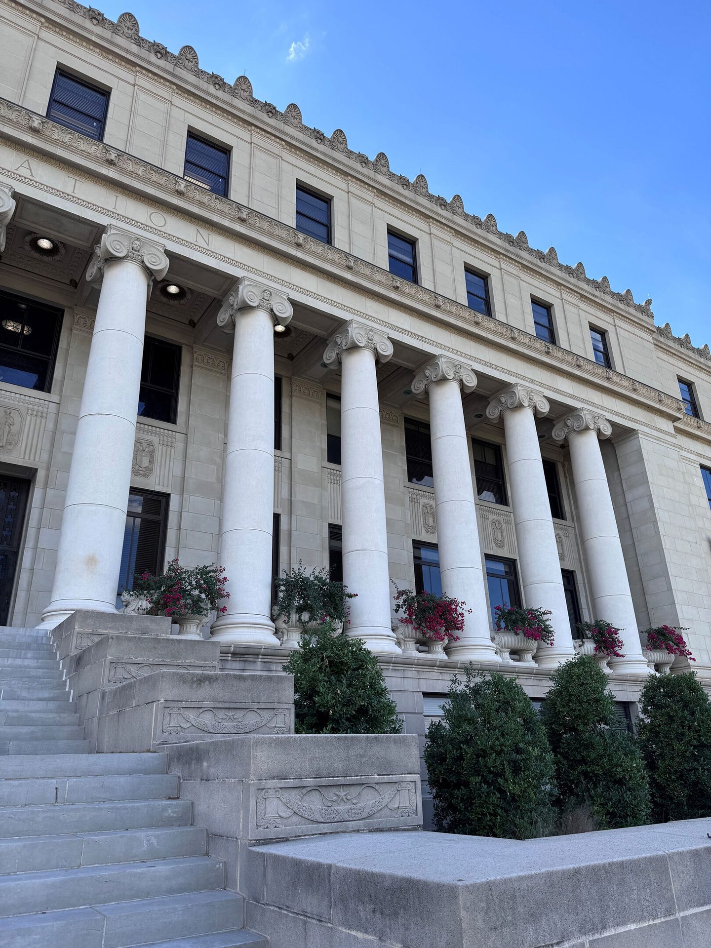 A grand, ornate stone building with tall white columns, a large staircase, and potted plants.