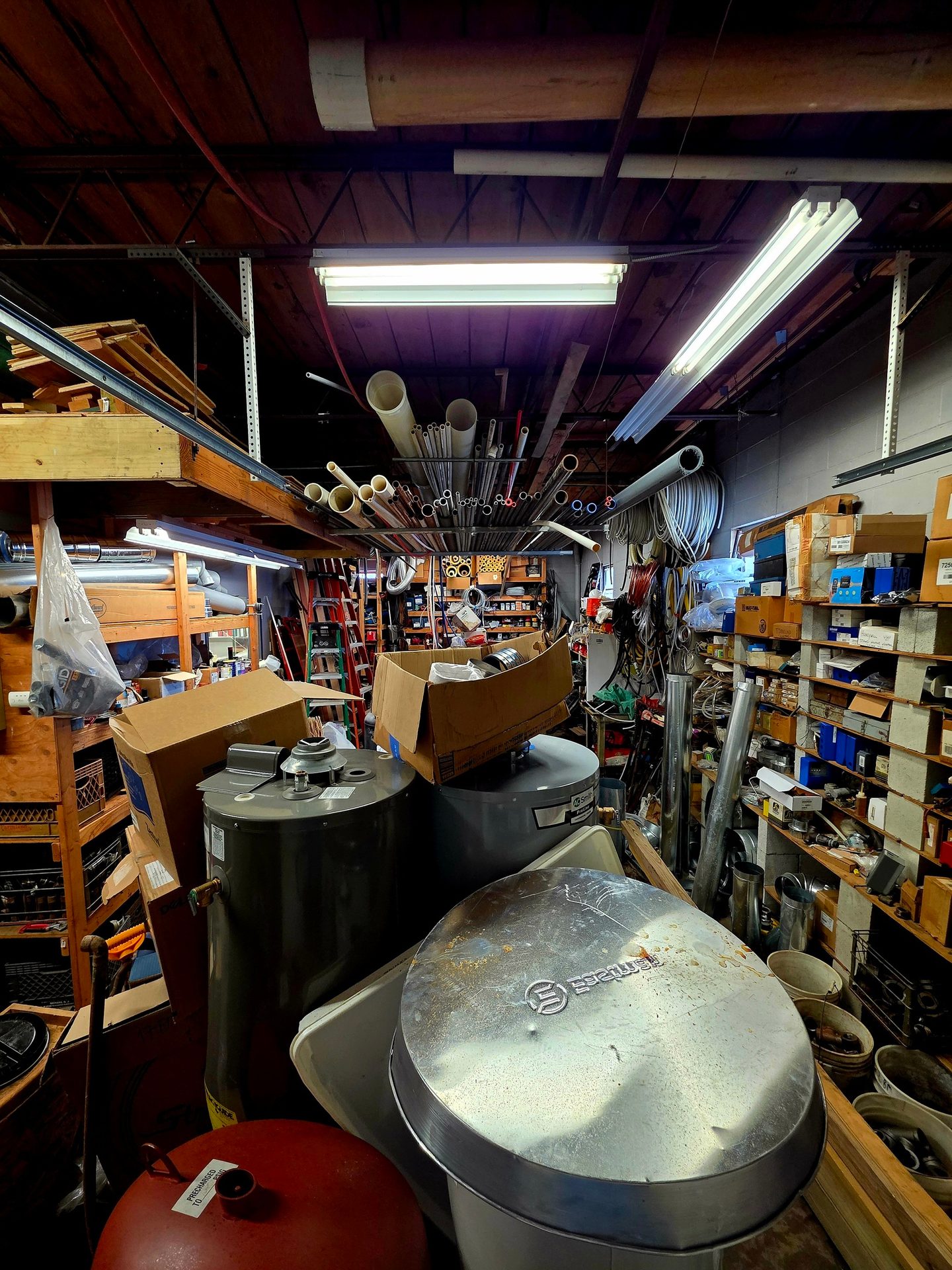 A cluttered storage room or warehouse full of plumbing supplies, pipes, water heaters, and shelving.
