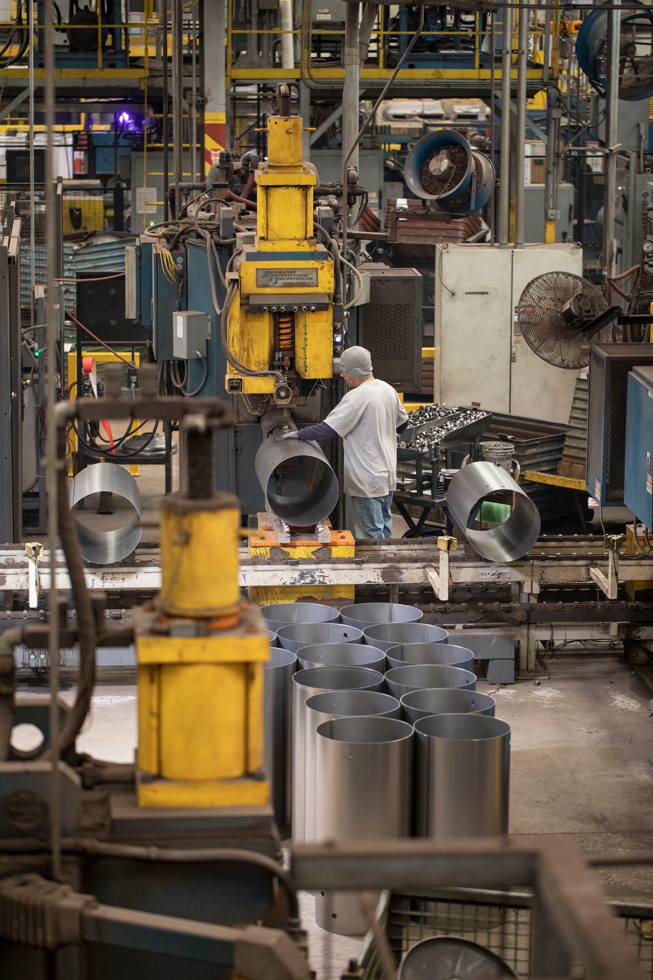 A worker operates industrial machinery to shape metal cylinders in a bustling factory setting.