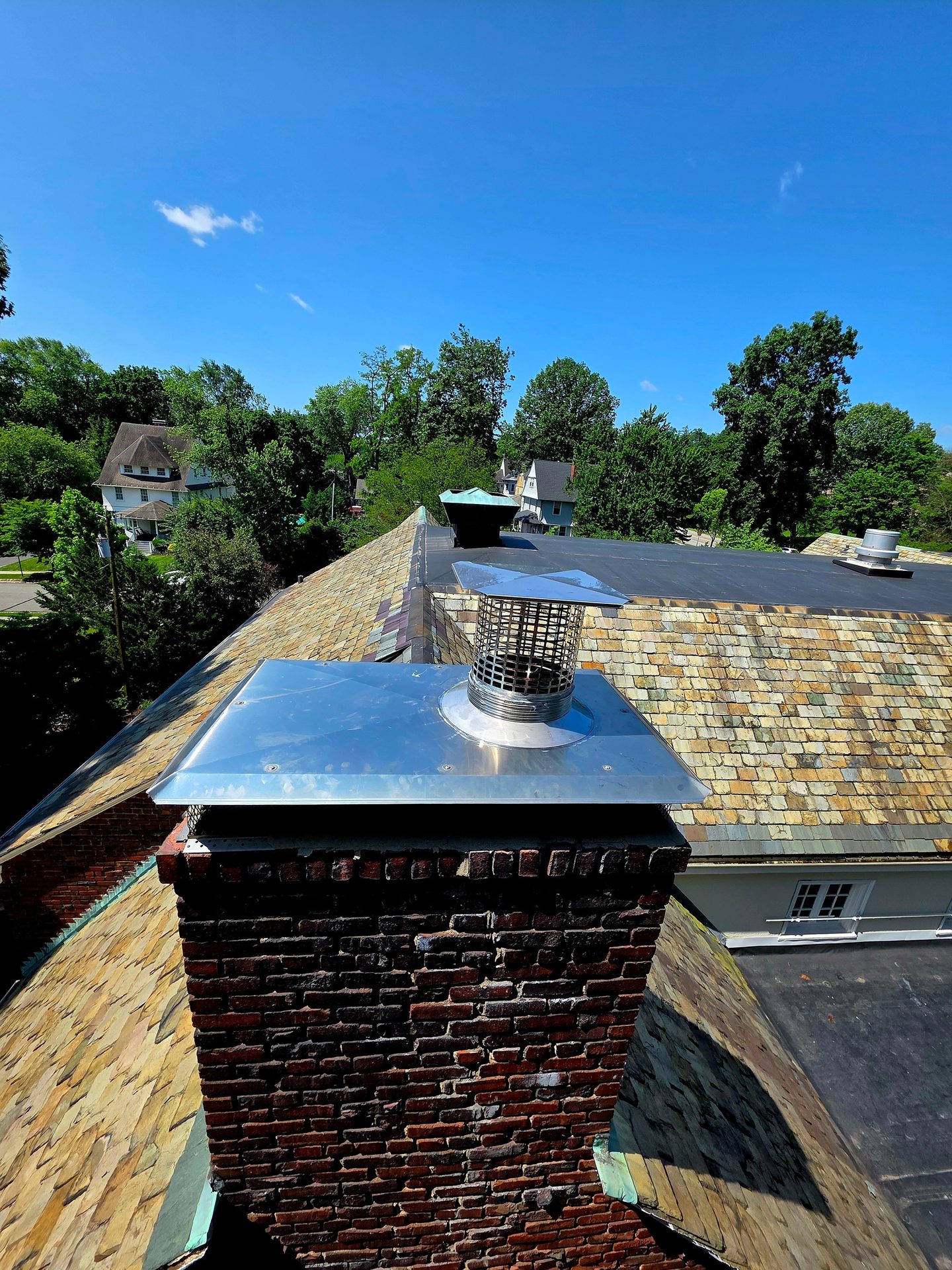 Close-up of a brick chimney with metal cap on a colorful slate roof, with trees and sky in background.