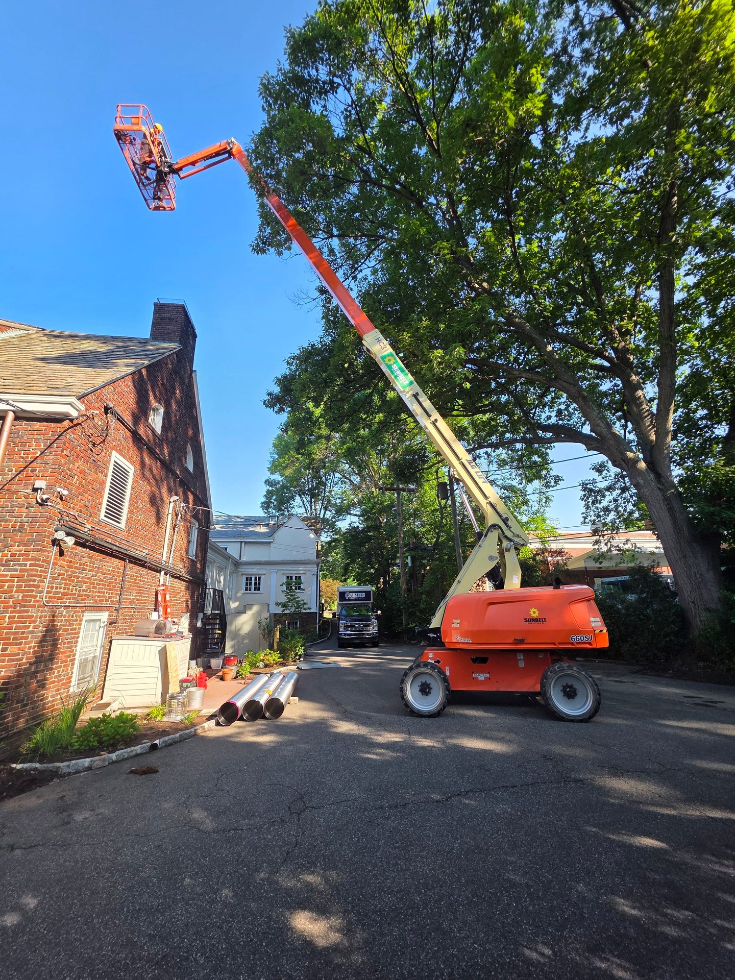 An orange boom lift extends high, carrying a worker who is trimming a tree next to a brick building.