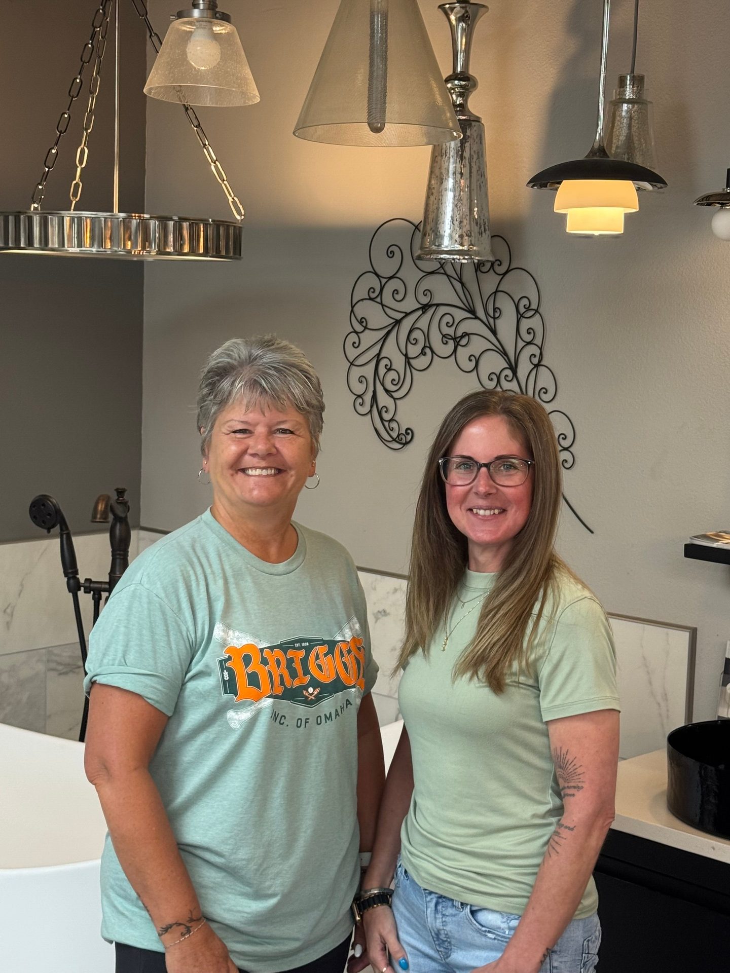 Two smiling women pose in a bright showroom featuring light fixtures and bath elements.