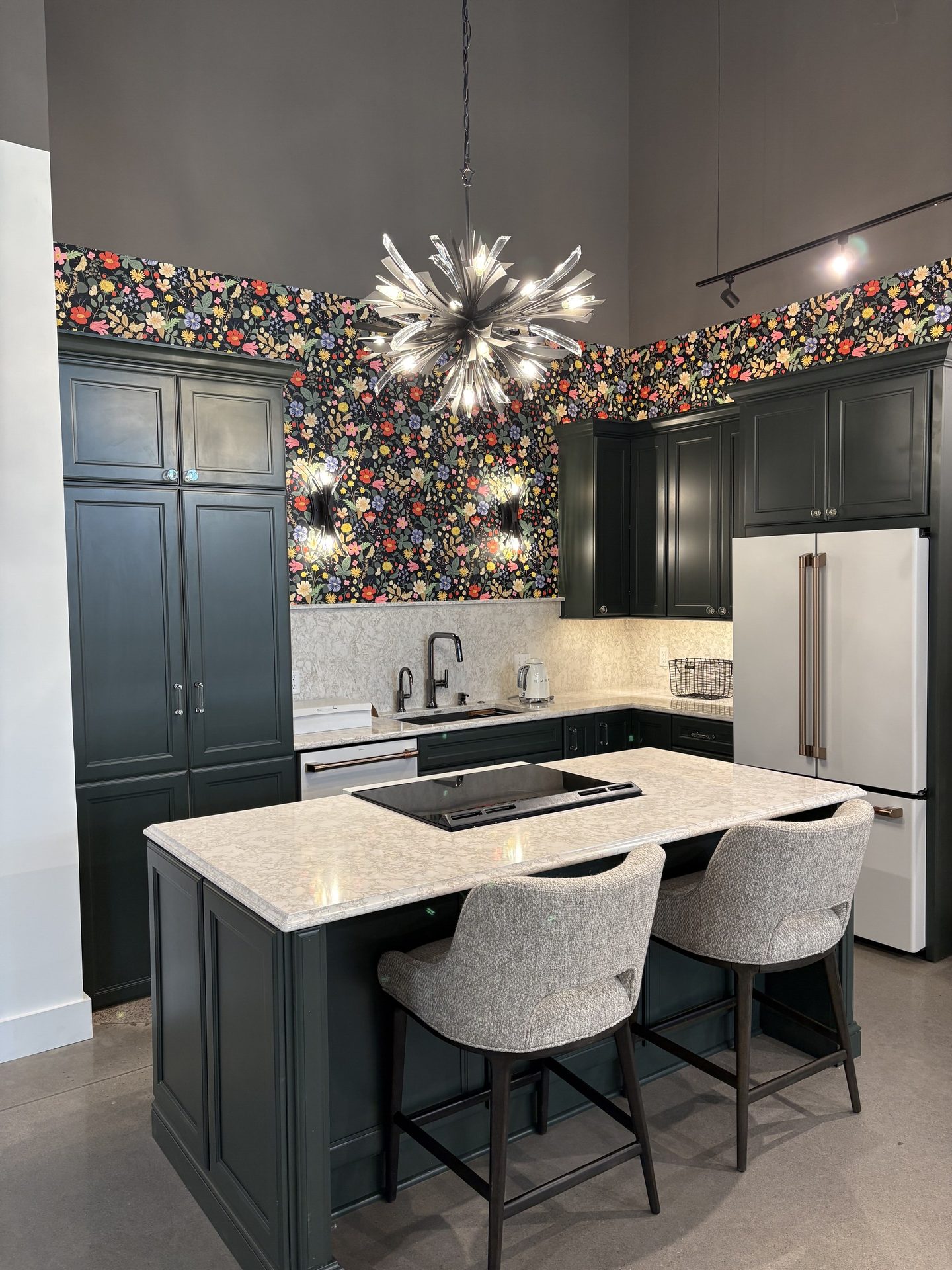 Kitchen with dark green cabinets, colorful floral wallpaper, light island, white fridge, and a sputnik chandelier.