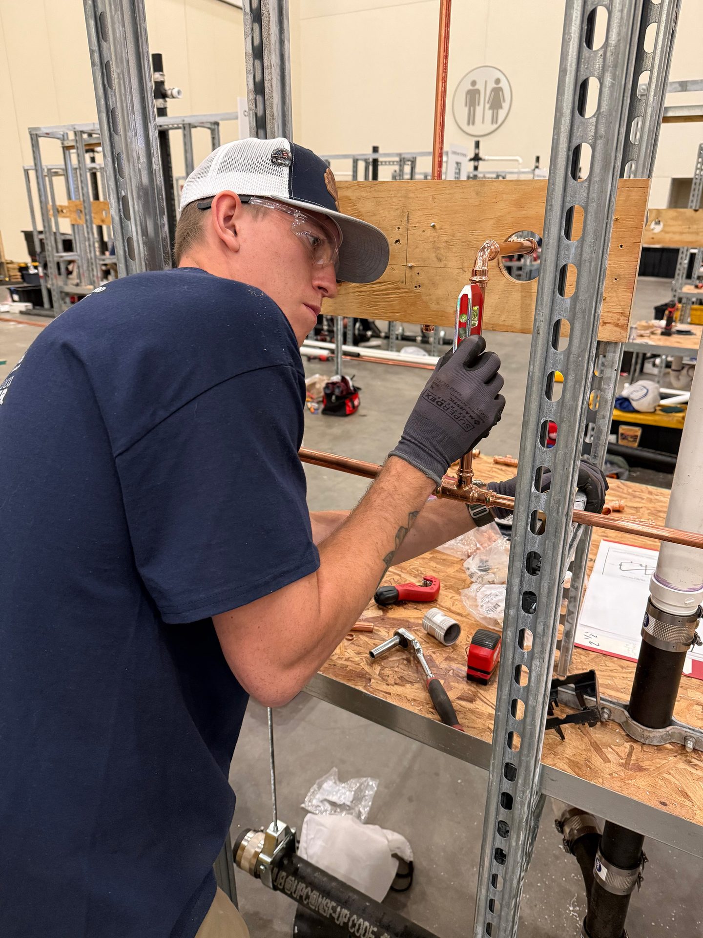 A man in safety glasses and a hat installs copper pipes, using a level, in a workshop.