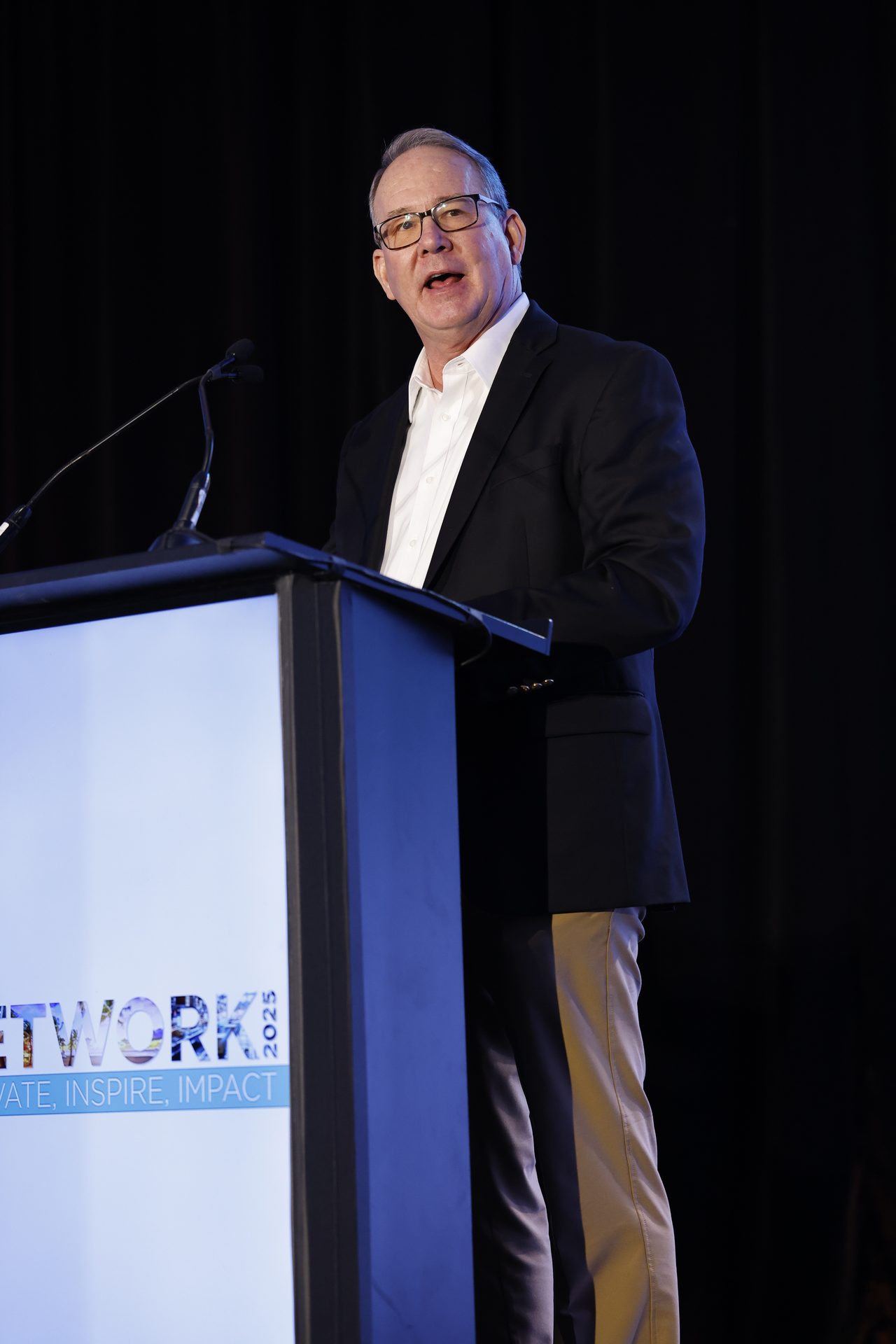 A man in glasses and a suit jacket speaks at a podium with microphones.