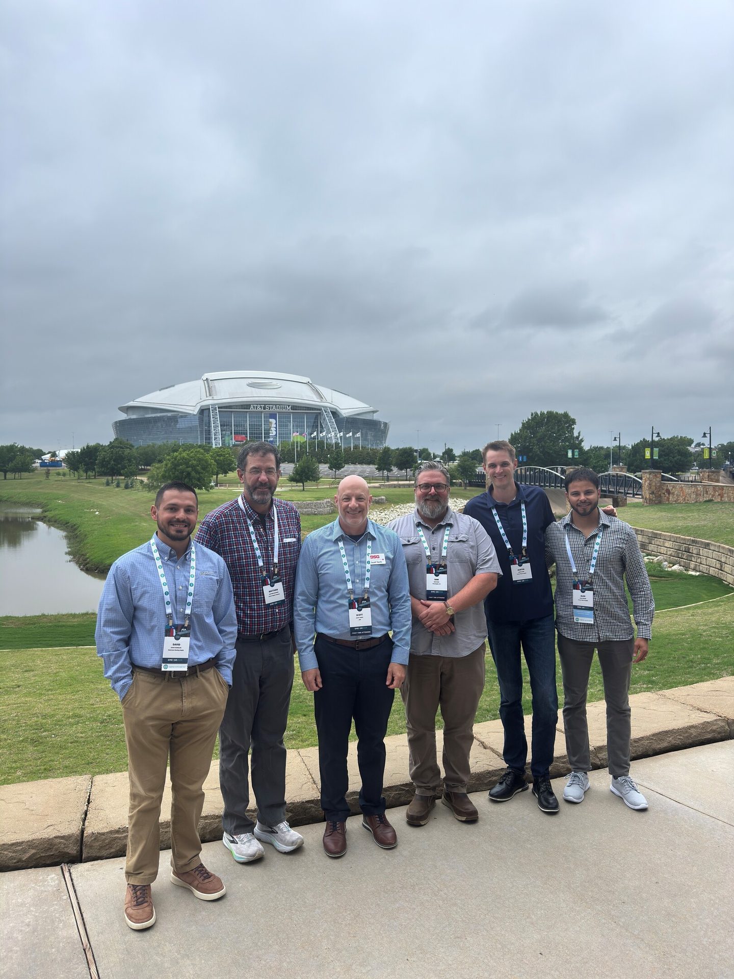 Six men stand in business casual attire with lanyards in front of AT&T Stadium under a cloudy sky.