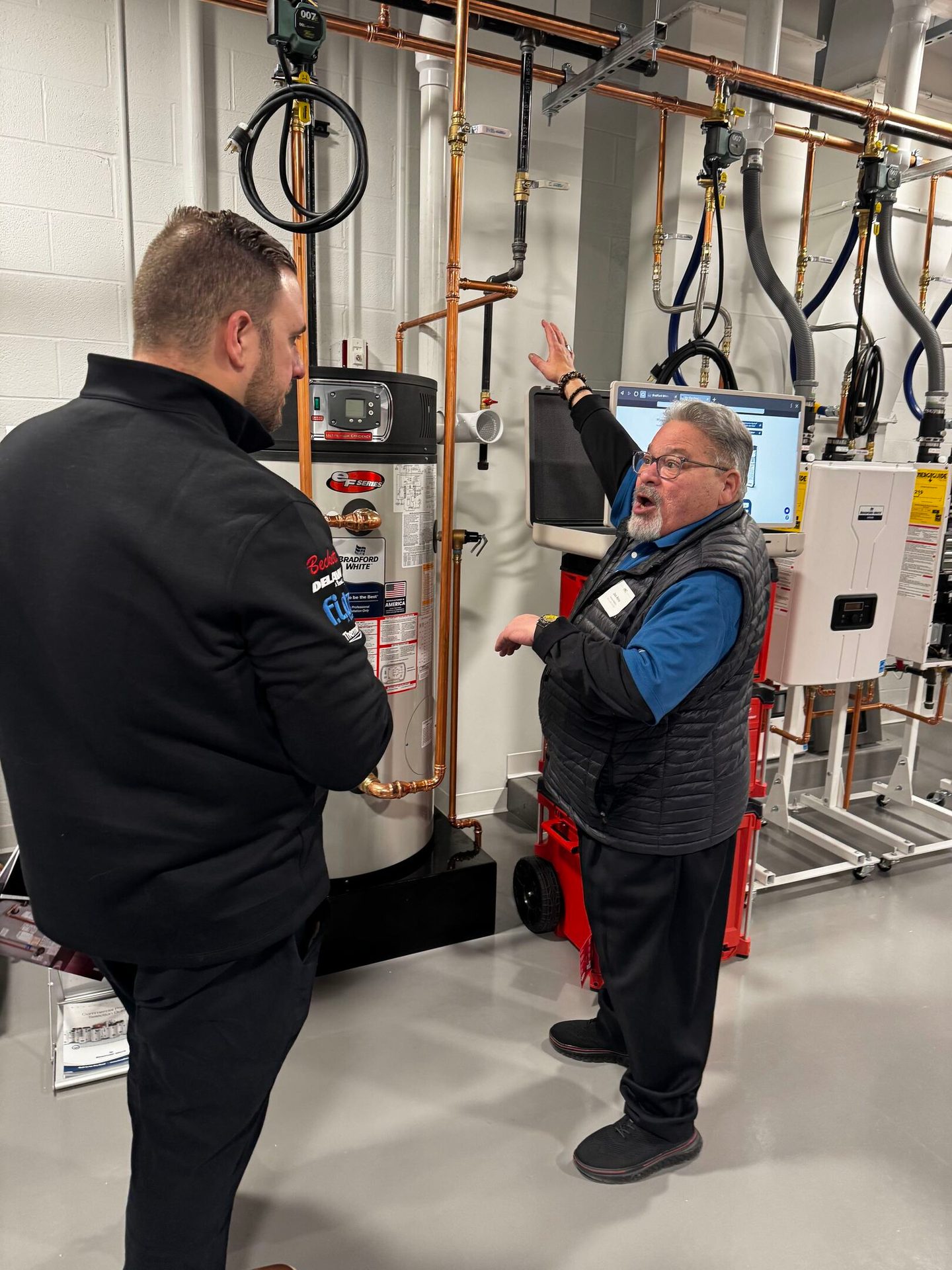 Two men discuss a complex plumbing system with a water heater, one gesturing to components.