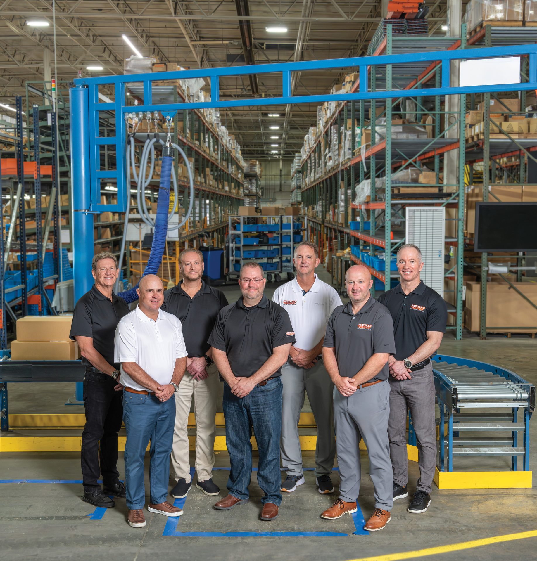 Seven men pose in a warehouse with a conveyor belt, industrial equipment, and tall racks.