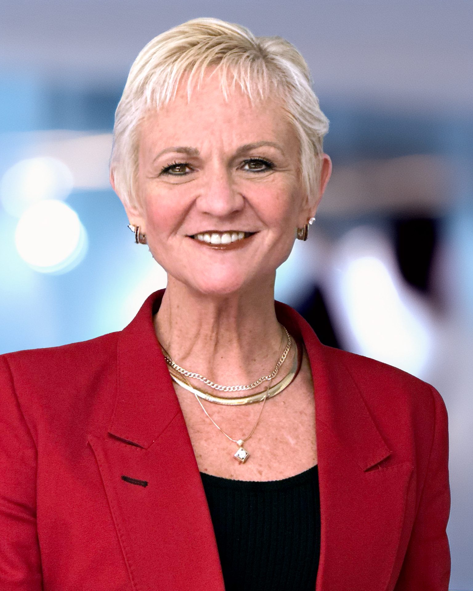 A smiling, blonde woman with short hair in a red blazer and gold jewelry.