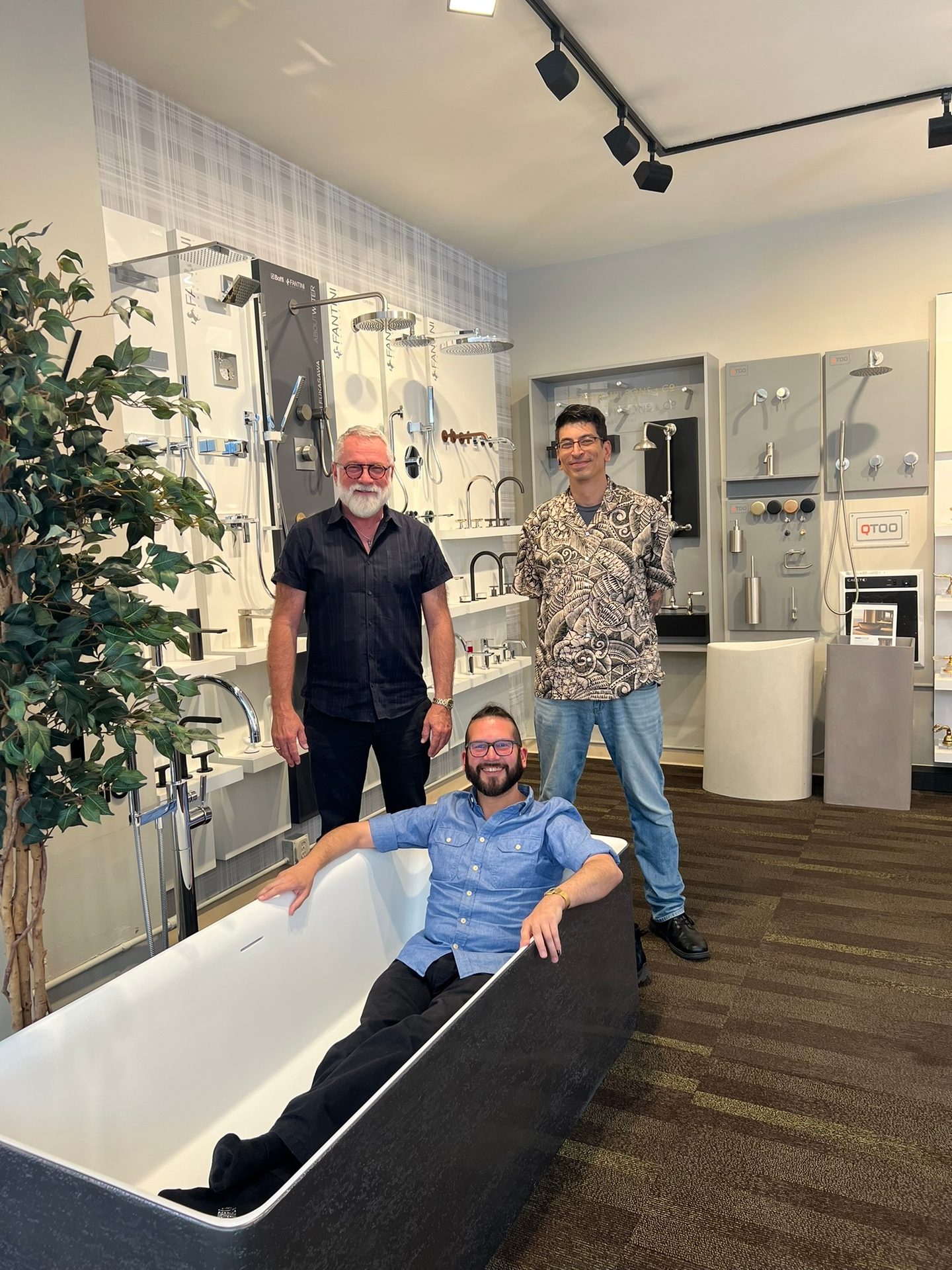 Three men in a bathroom showroom, one sitting in a bathtub, surrounded by fixtures.