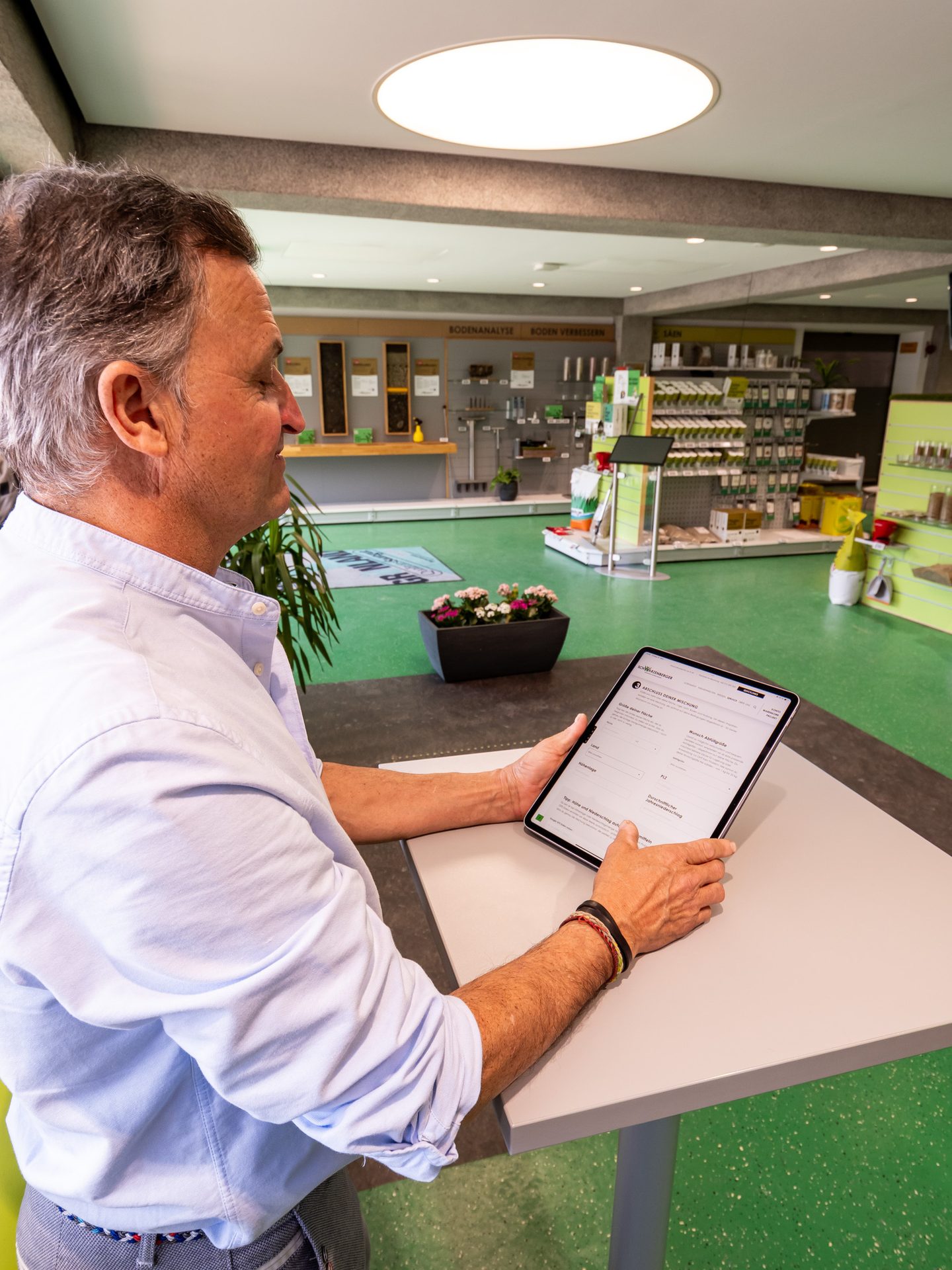 Man in a light blue shirt uses a tablet in a garden supply store with shelves of products in the background.