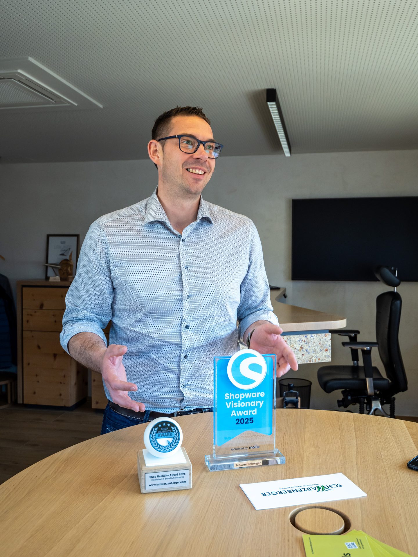 A man in glasses and a light blue shirt stands smiling behind a table with two awards, gesturing with his hands.