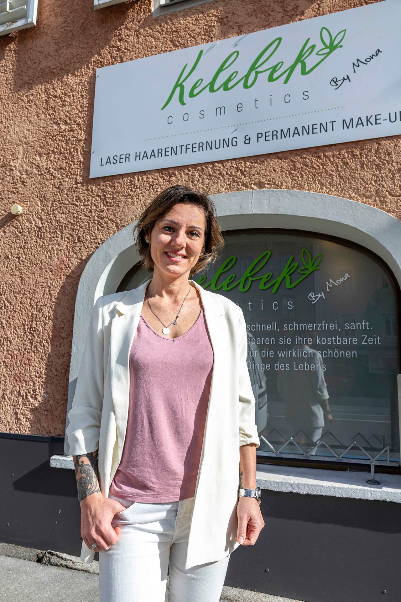 A smiling woman in a white blazer and pink top stands in front of a beauty salon sign for Kelebek Cosmetics.