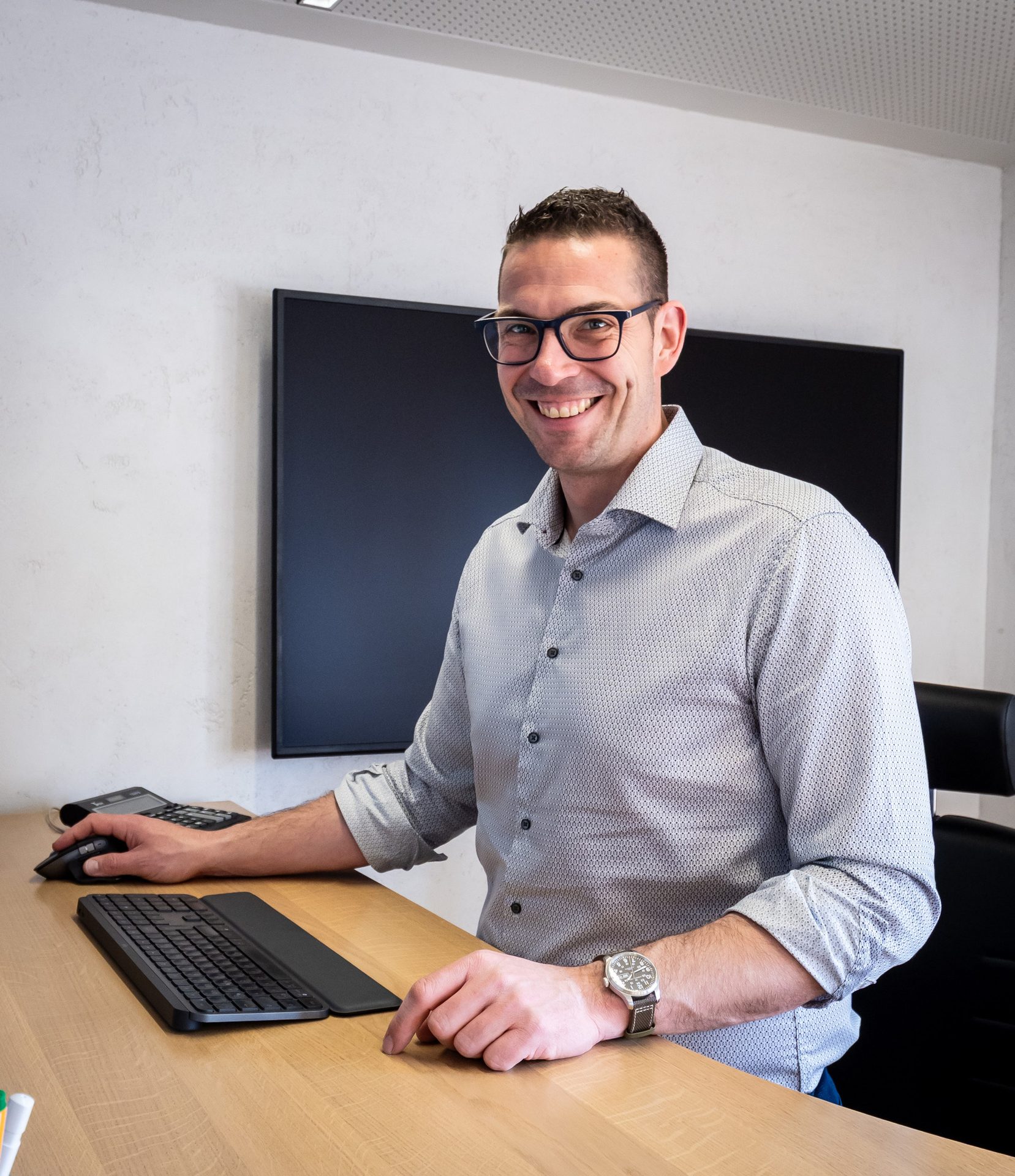 A smiling man wearing glasses and a patterned shirt works at a desk with a keyboard and mouse.
