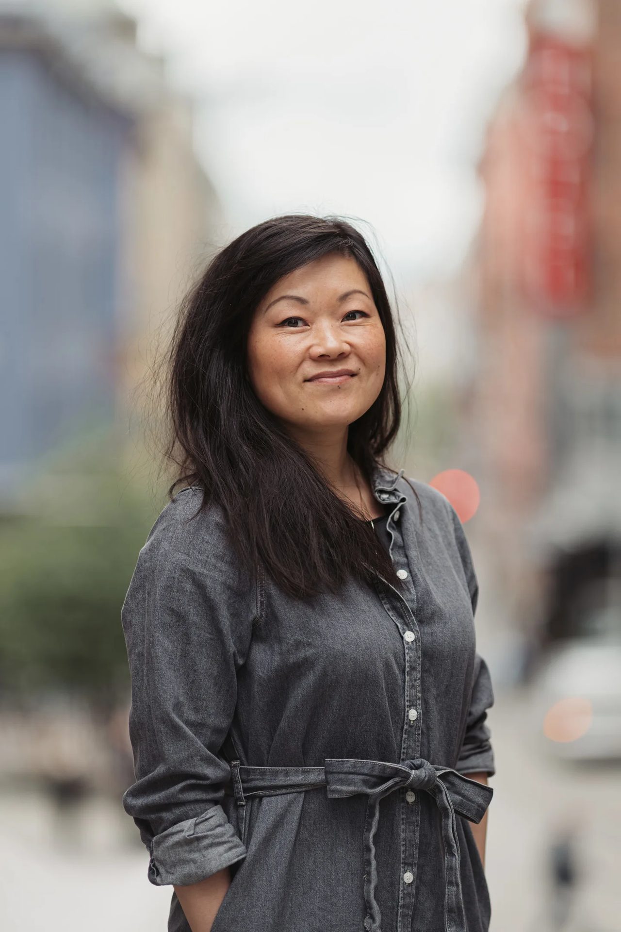 Smiling woman with dark hair in a gray denim shirtdress stands outdoors.