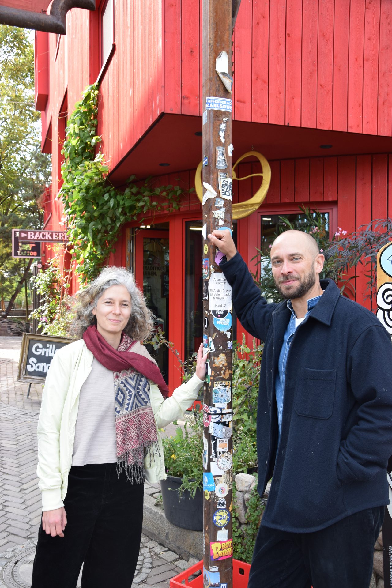 Man and woman lean on a sticker-covered pole outside a red building with bakery and tattoo signs.