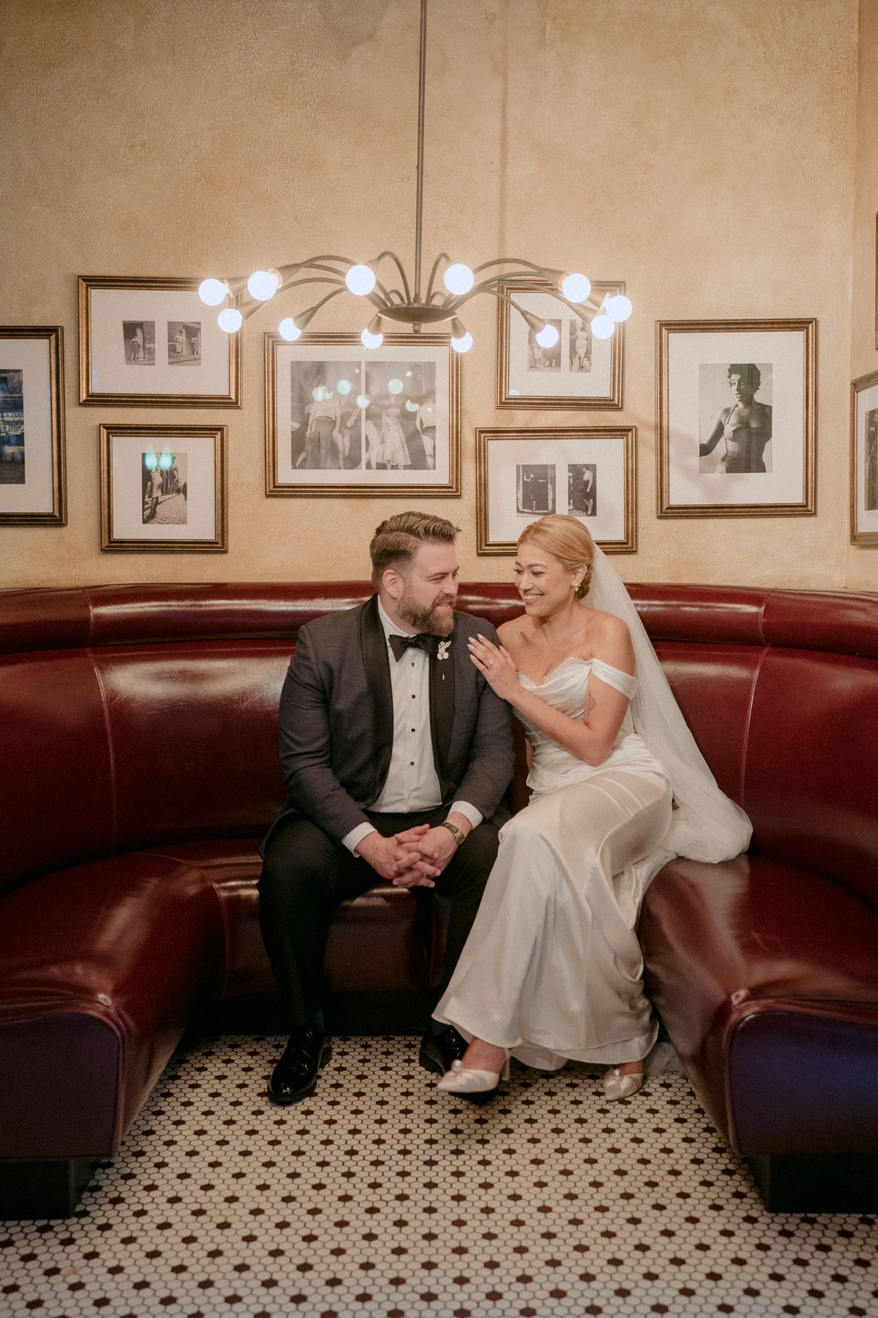 A happy wedding couple in formal attire sits on a red booth, gazing lovingly at each other.