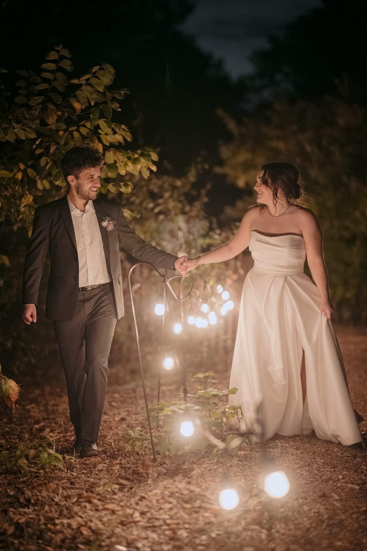 A smiling couple in formal wear holding hands on a path at night, illuminated by string lights.